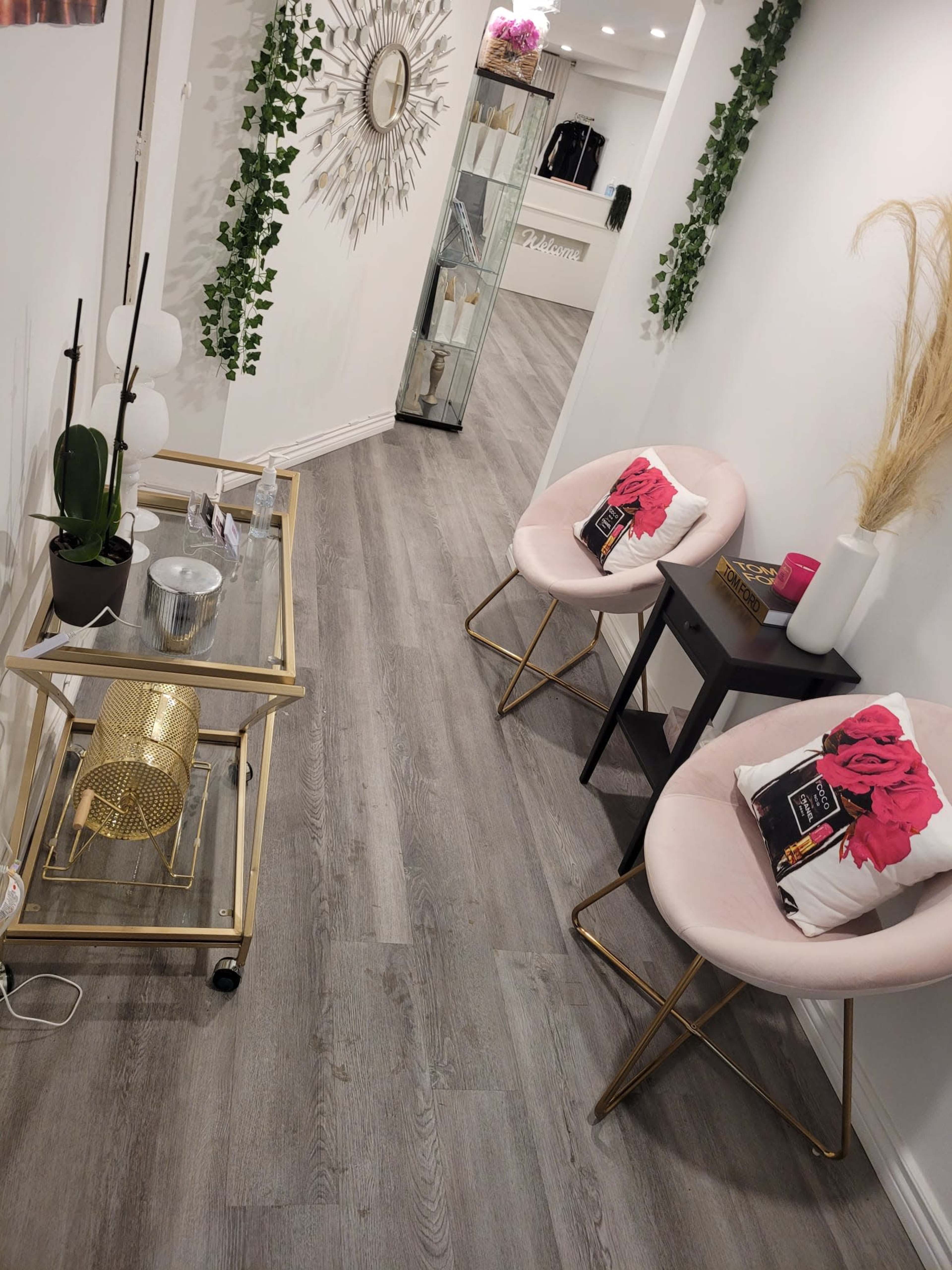 A narrow, modern hallway with light gray wooden flooring, featuring two pink chairs with floral cushions, a glass cart, and decorative plants on the wall.