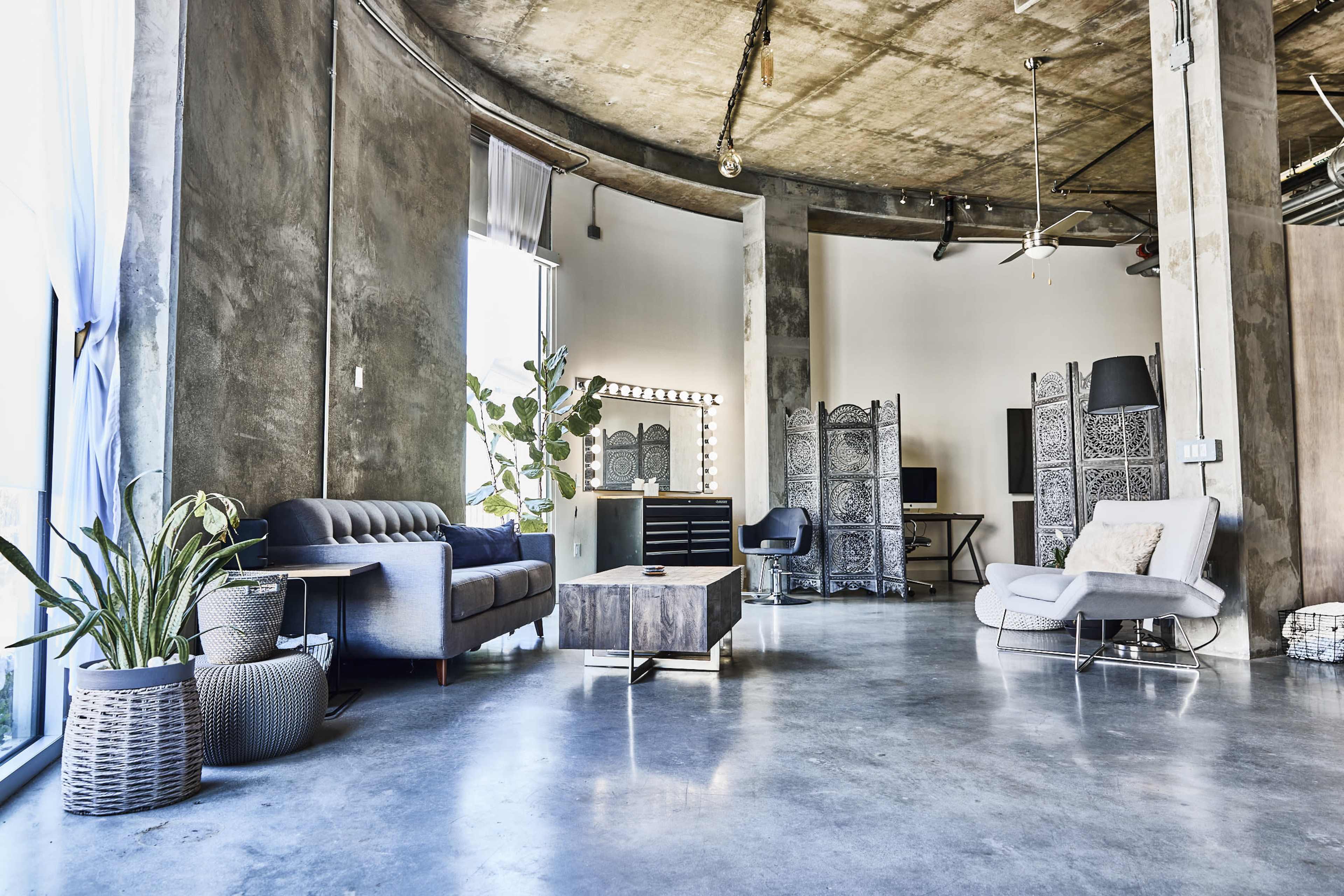 The image shows a spacious industrial-style living area featuring a concrete floor, large windows providing natural light, a gray sofa, a coffee table, indoor plants, and decorative room dividers.