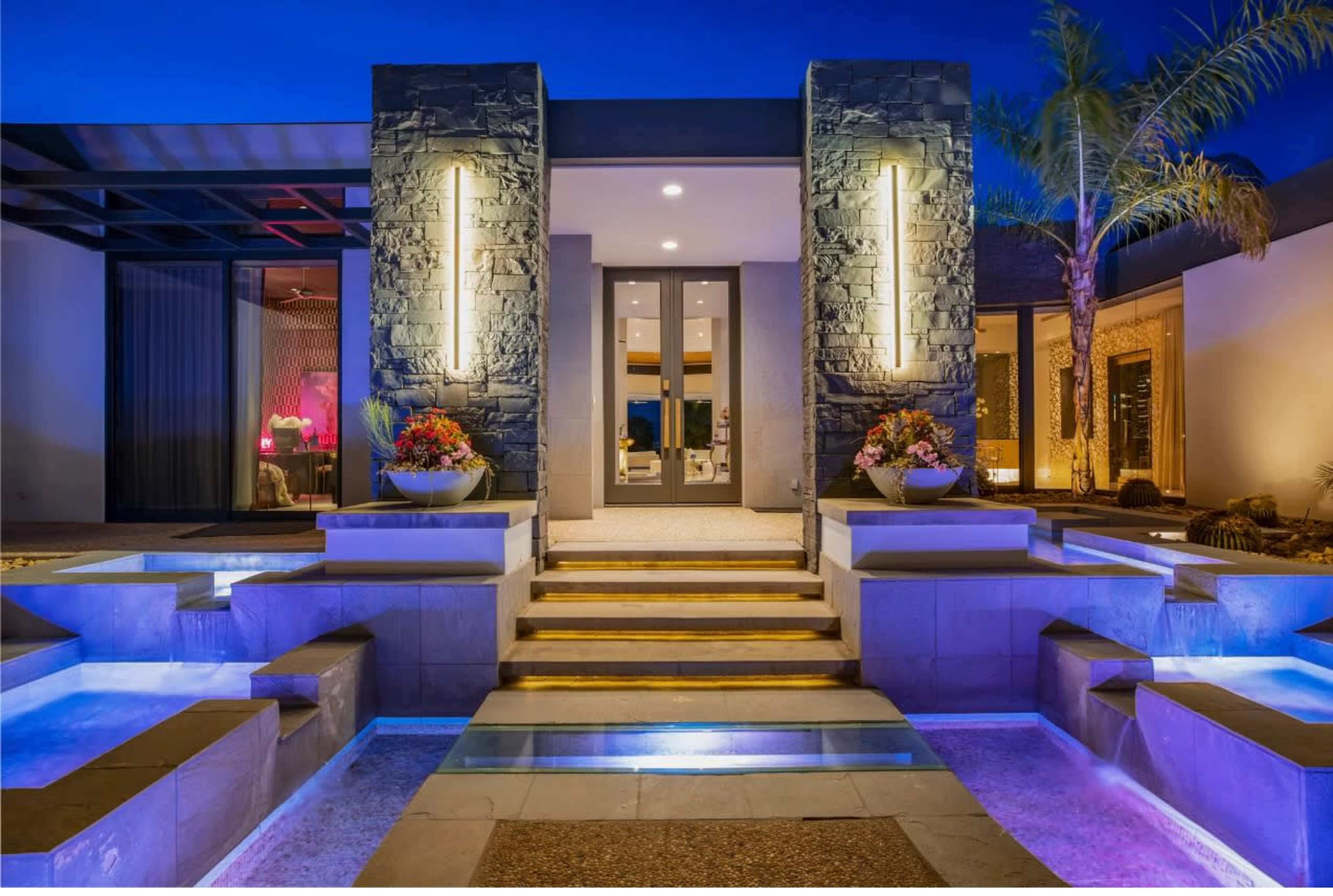 The image showcases a modern home entrance with stone accents, illuminated by two vertical lights, flanked by decorative planters and a water feature.