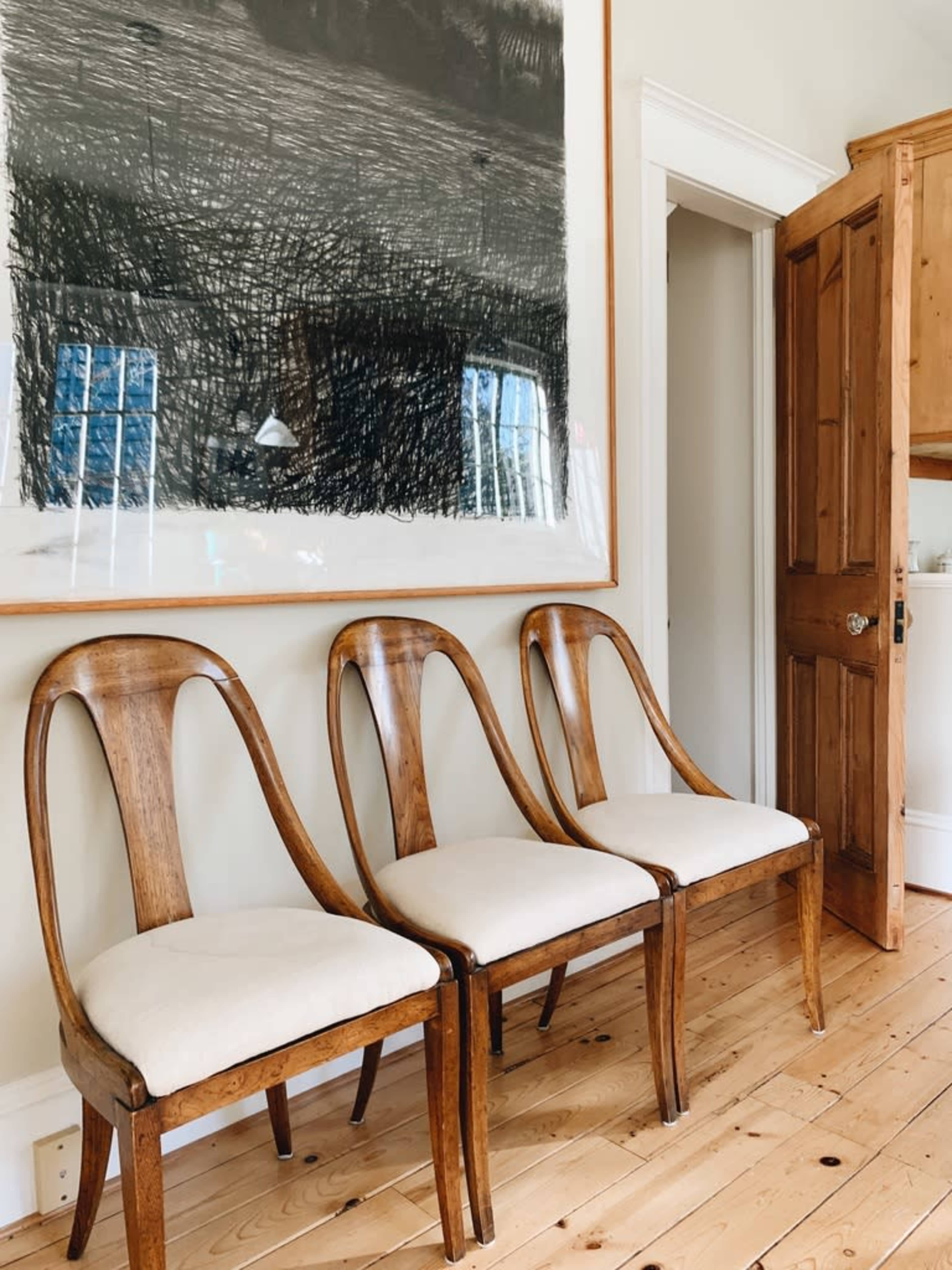 Three wooden chairs with upholstered seats are lined up against a wall beneath a large artwork, while a wooden door stands partially open in the background.