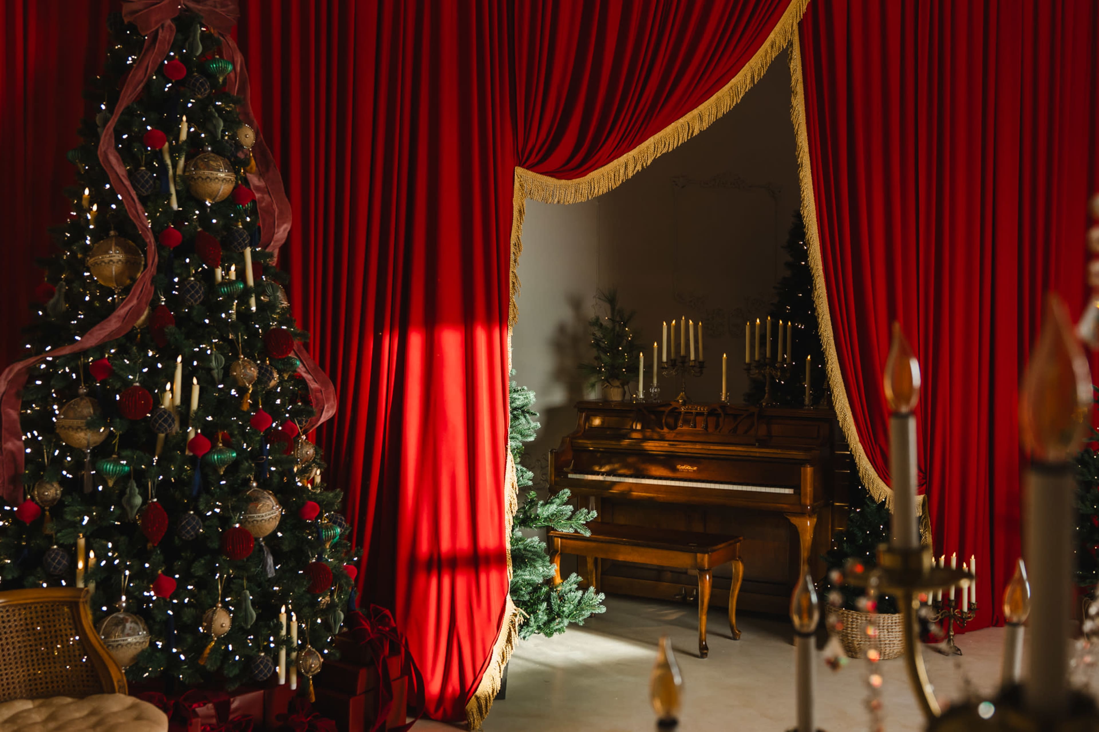 A beautifully decorated room features a grand piano flanked by two Christmas trees, surrounded by vibrant red curtains and soft candlelight.