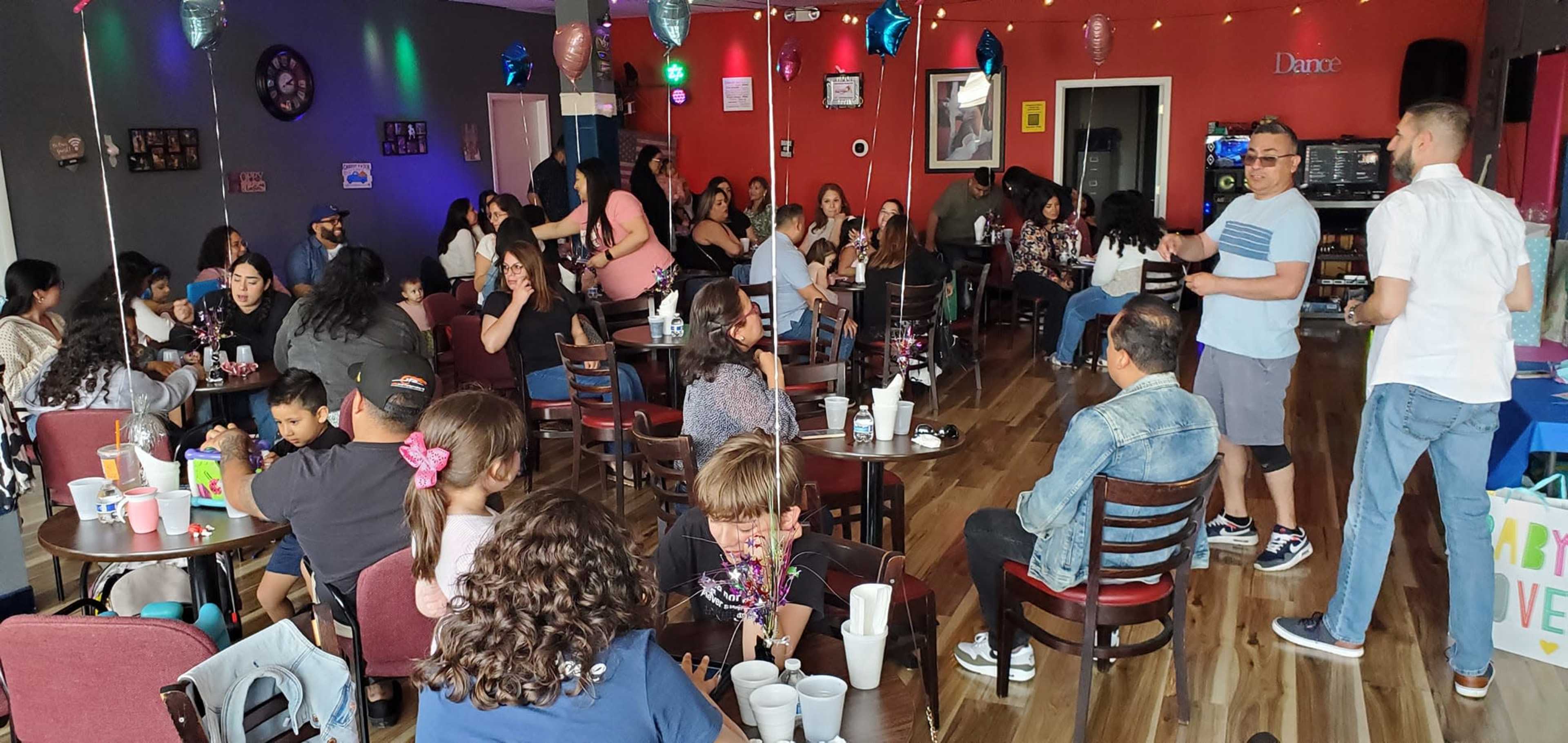 The image shows a lively café filled with groups of people socializing at tables, with decorations and balloons adding to the festive atmosphere.
