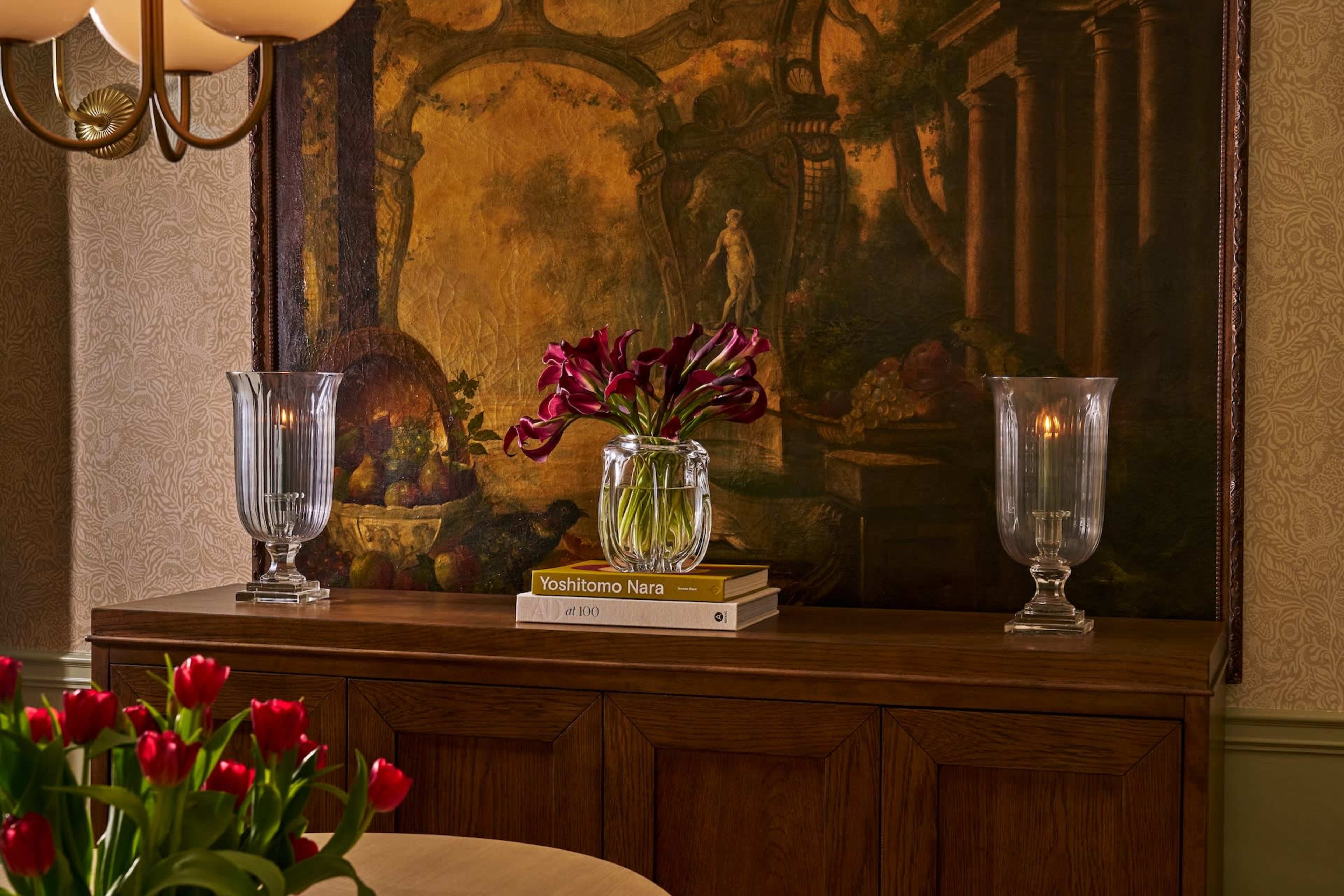 A wooden sideboard displays two glass vases containing tulips and a decorative book against a large, classical painting.