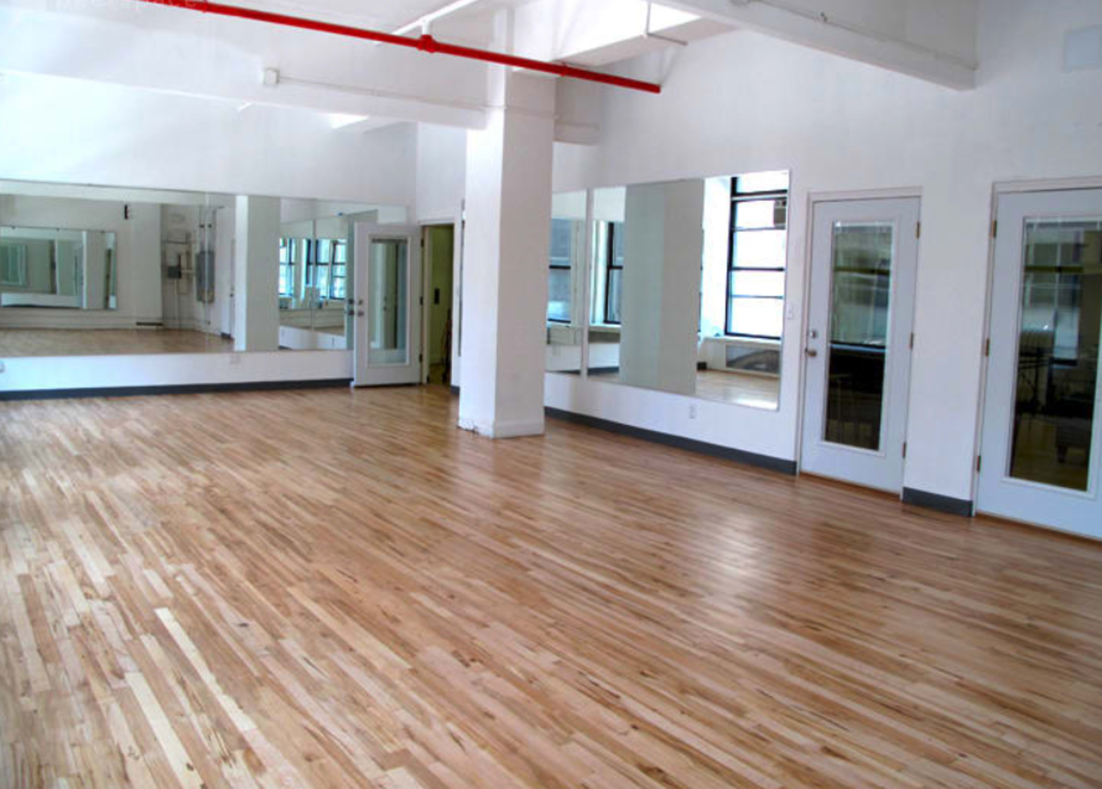 The image shows an empty room with polished hardwood floors, large mirrors on the walls, and two double doors leading to another area.