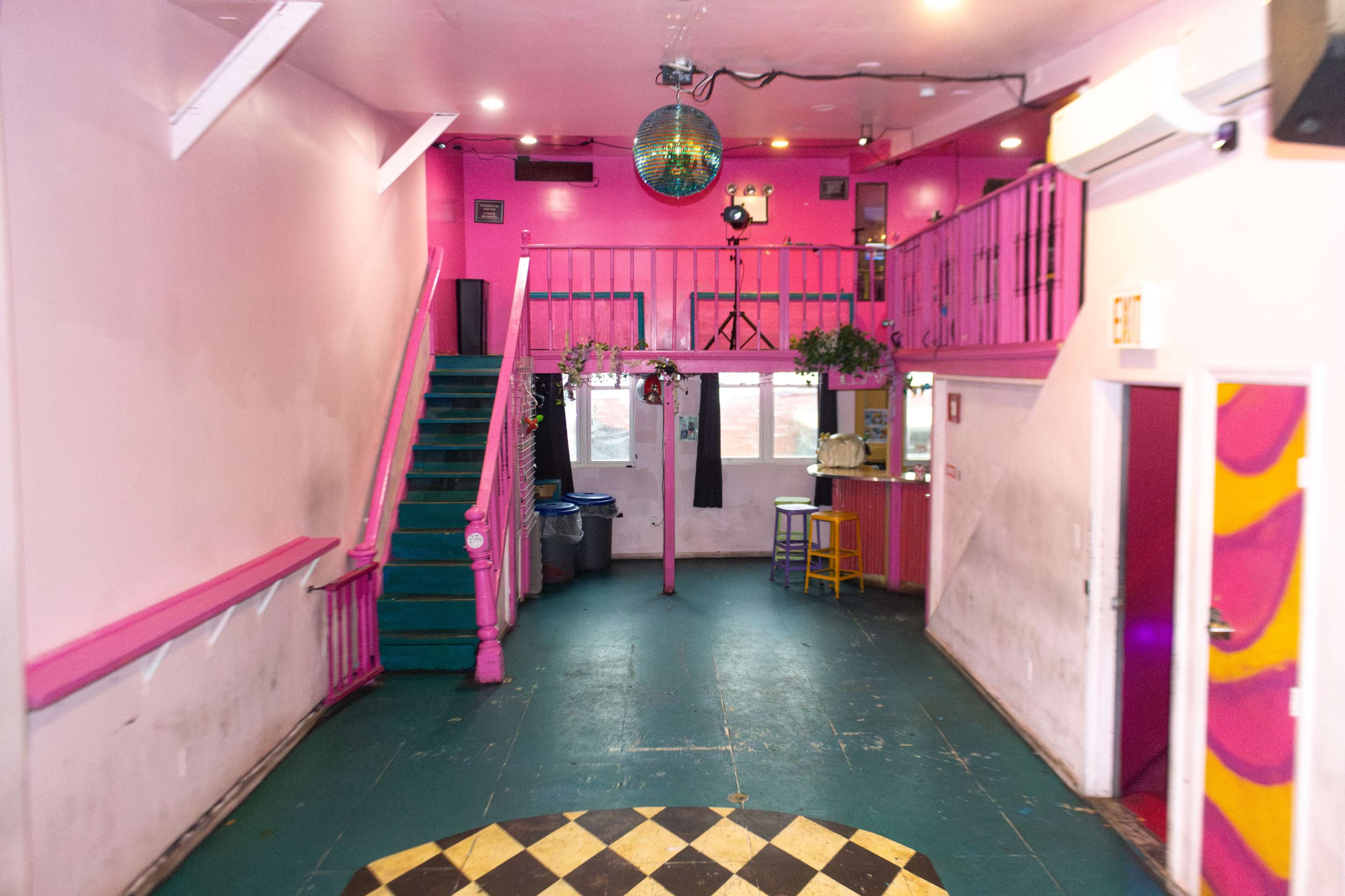 The image shows a vibrant interior space with pink walls, a disco ball hanging from the ceiling, and a staircase leading to a mezzanine level.
