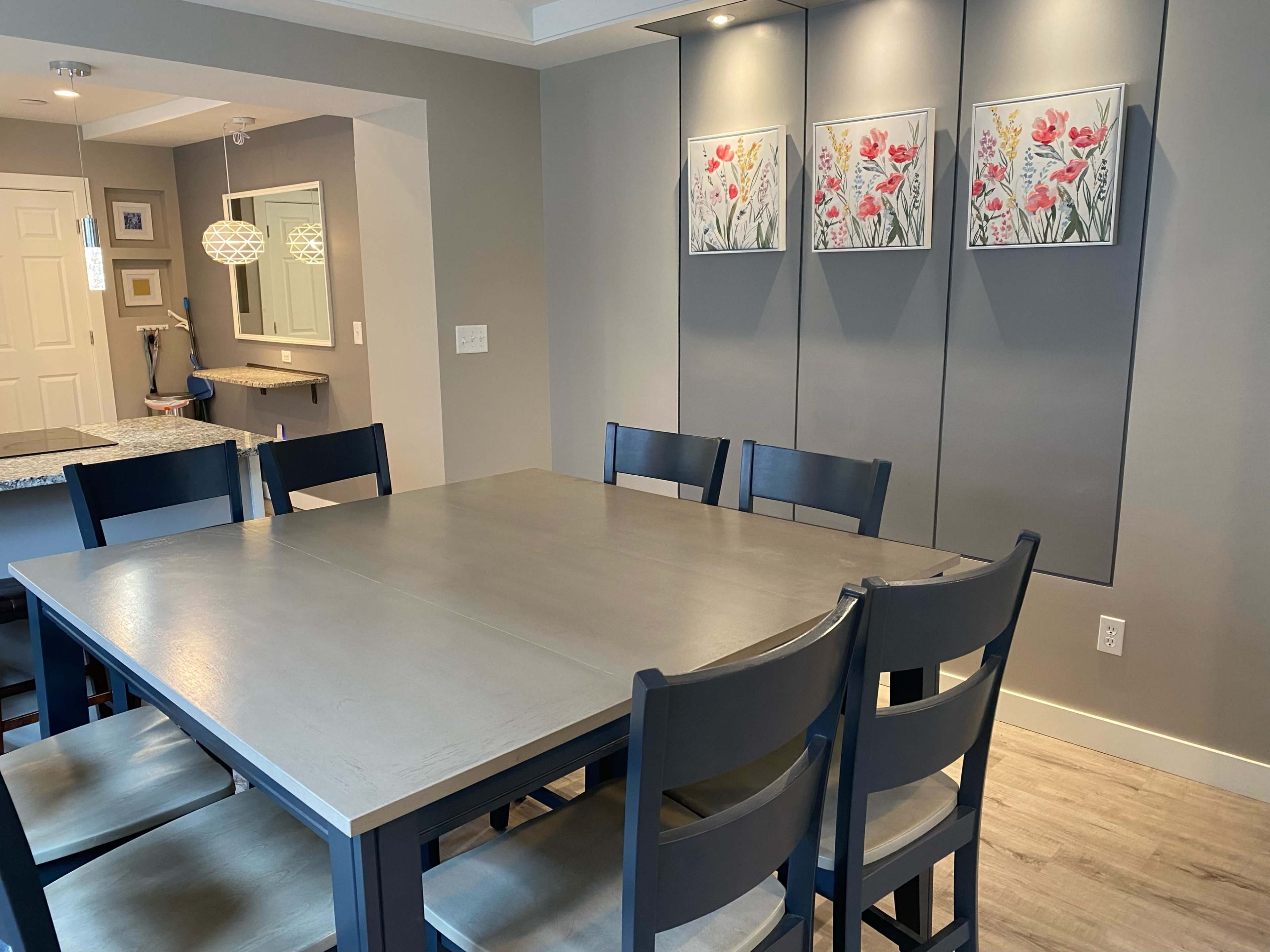 A large wooden dining table is surrounded by six black chairs in a well-lit room featuring floral artwork on the wall.