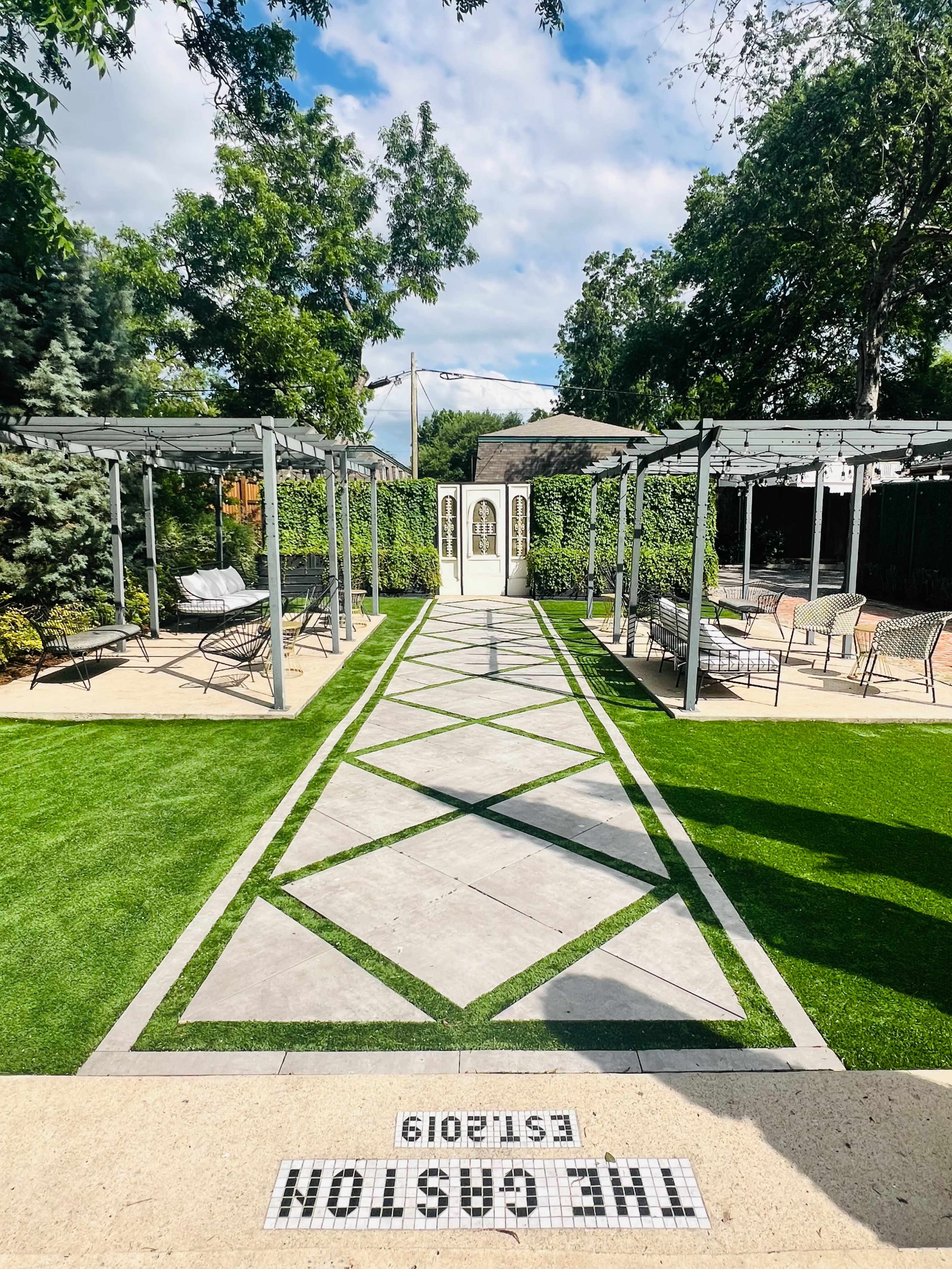 The image shows a landscaped garden with a pathway made of leaf-shaped concrete tiles bordered by greenery and modern pergolas.