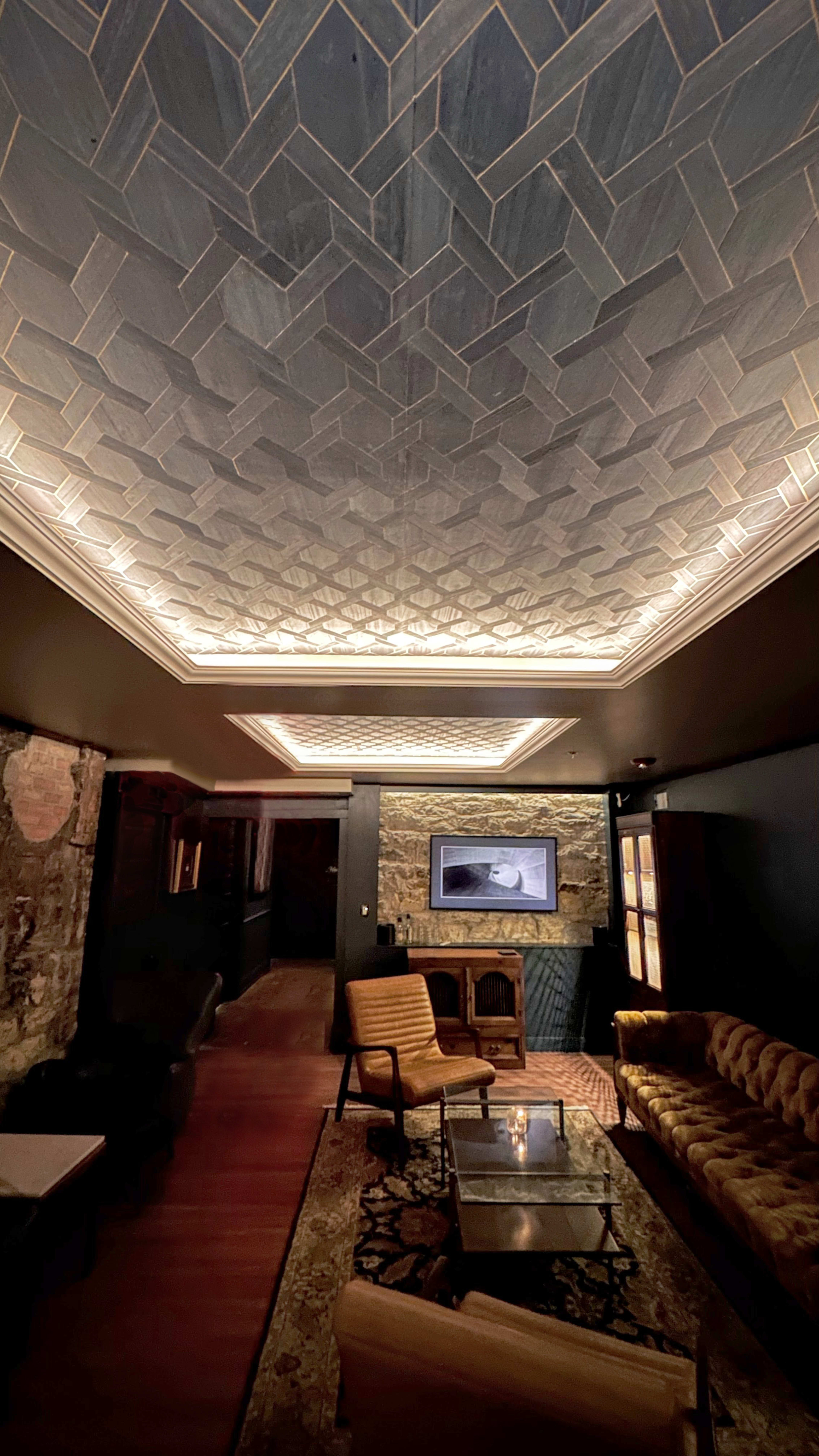 A dimly lit lounge with a patterned ceiling, exposed brick walls, and a vintage sofa, accompanied by modern lighting and a wall-mounted screen.