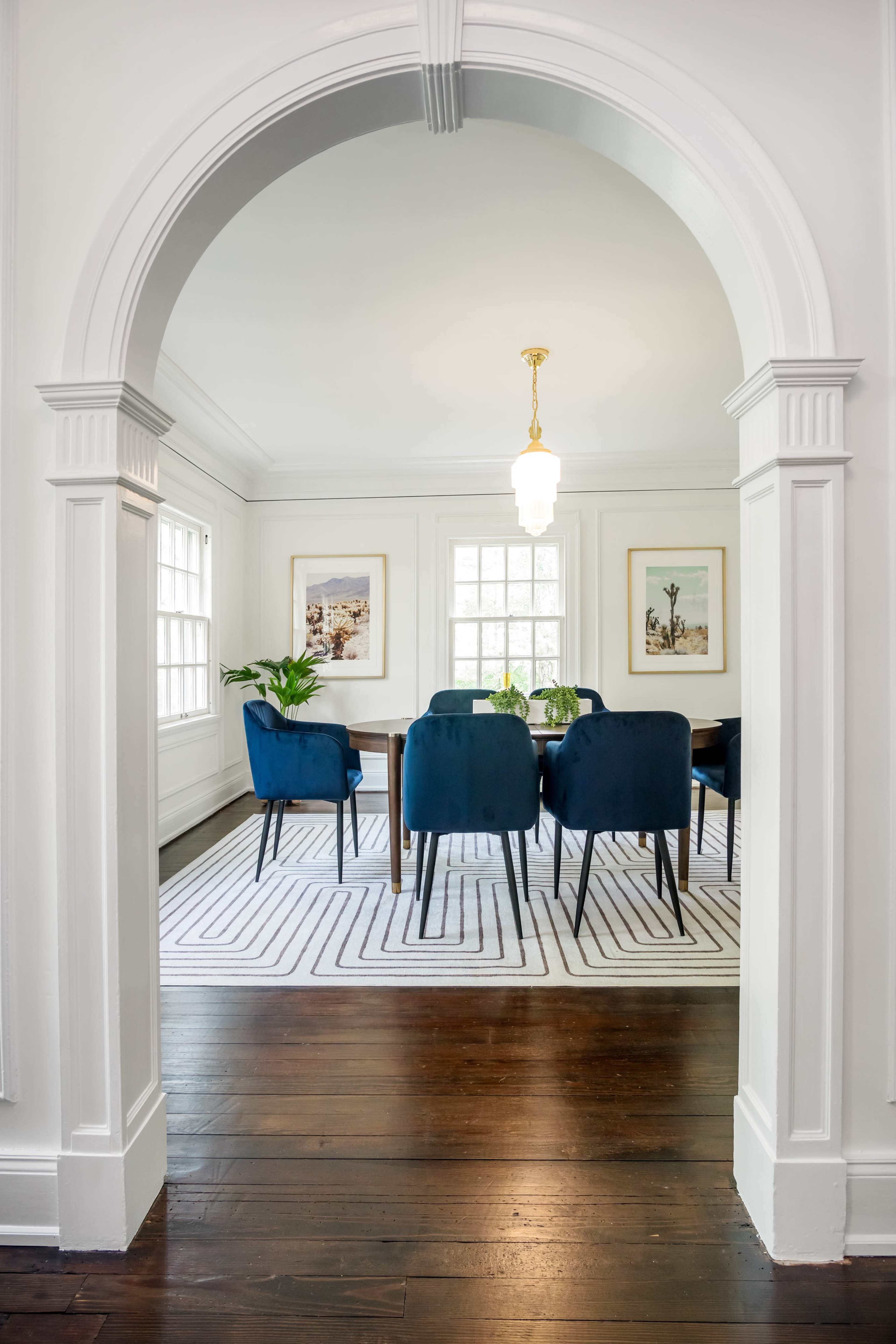 A dining room features a long table surrounded by dark blue chairs, viewed through an archway with a light fixture overhead.