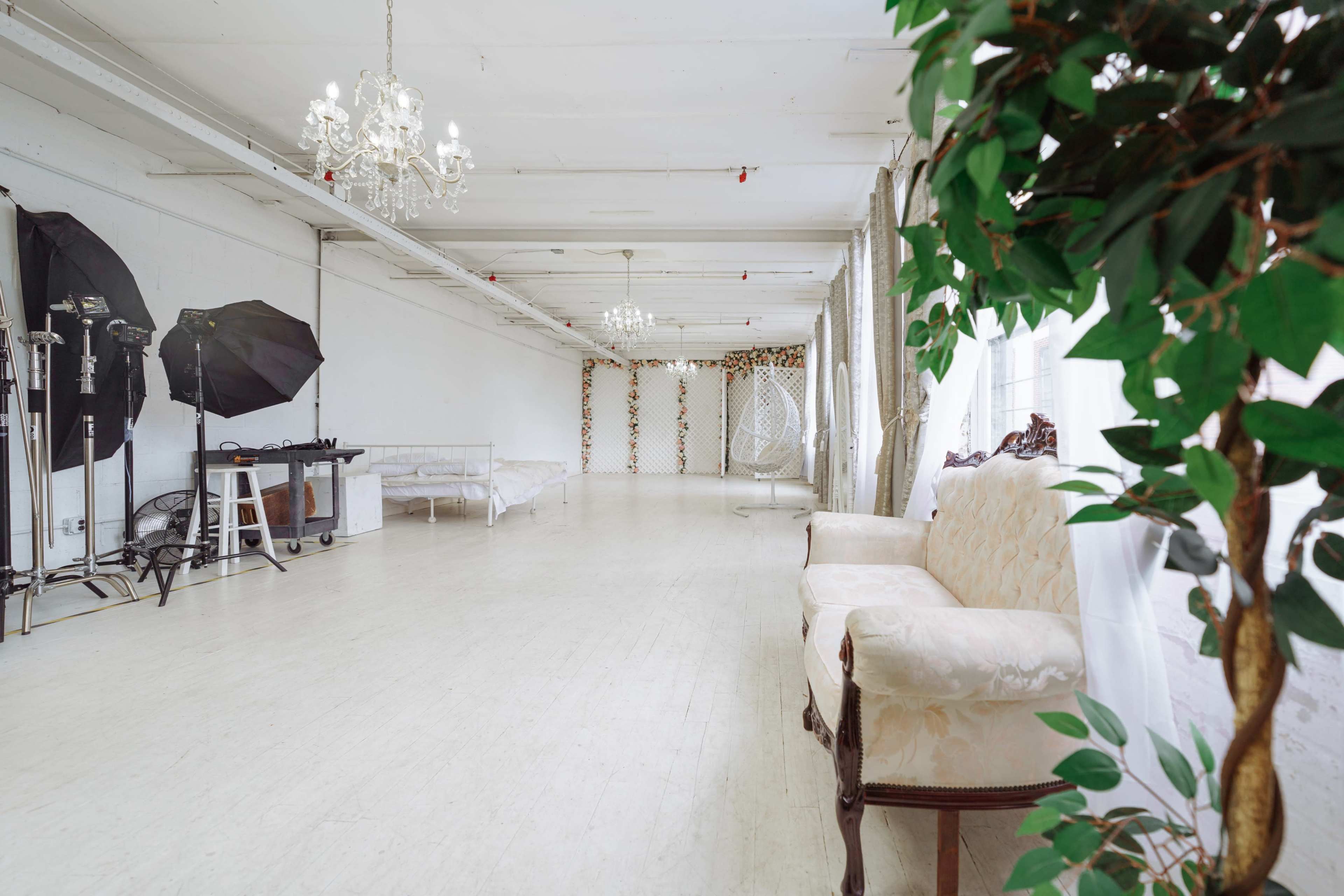 A spacious, brightly lit studio with a light-colored wooden floor, chandeliers, and a vintage sofa beside photography equipment and decorative elements.