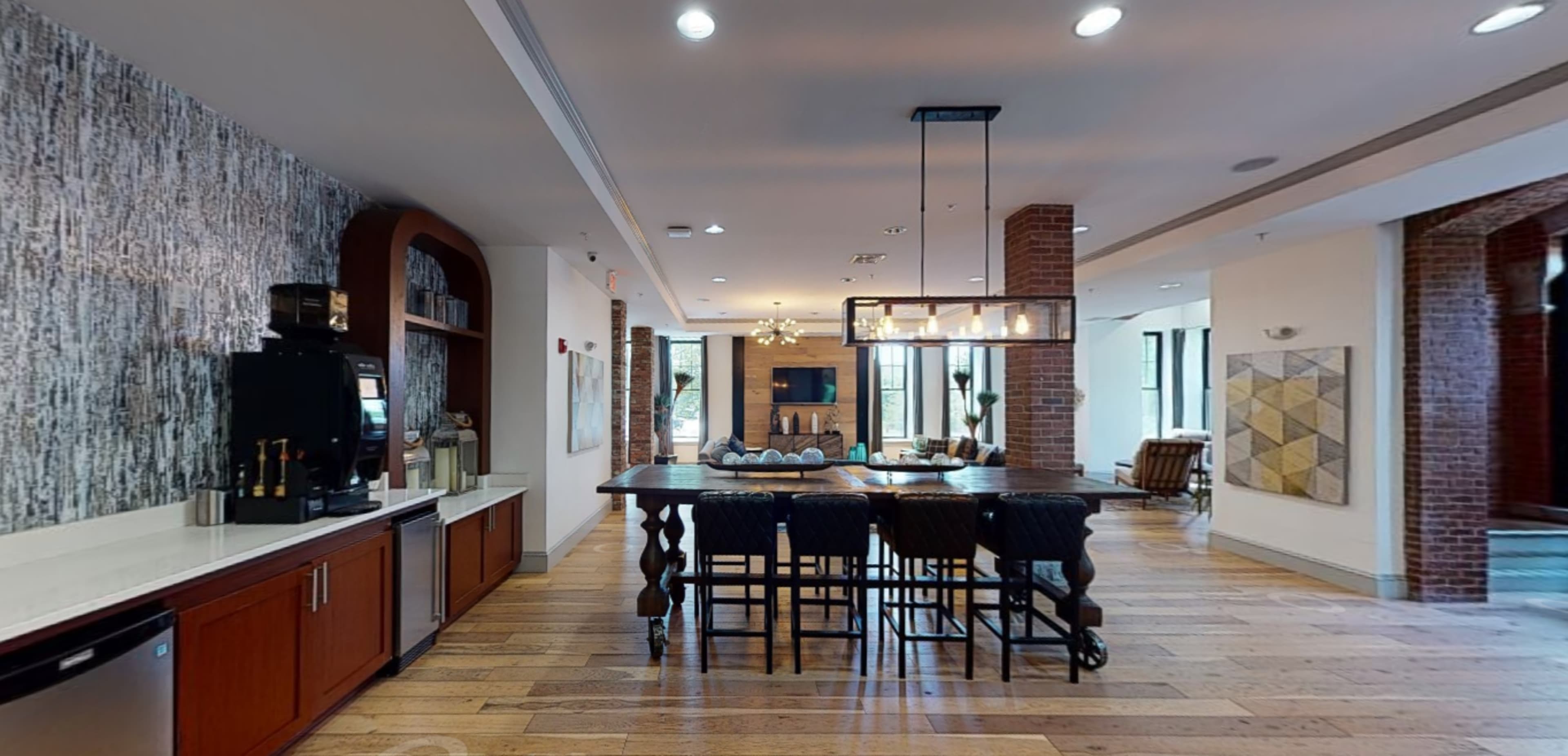 A modern interior space with a coffee station, a large dining table surrounded by chairs, and a seating area in the background.