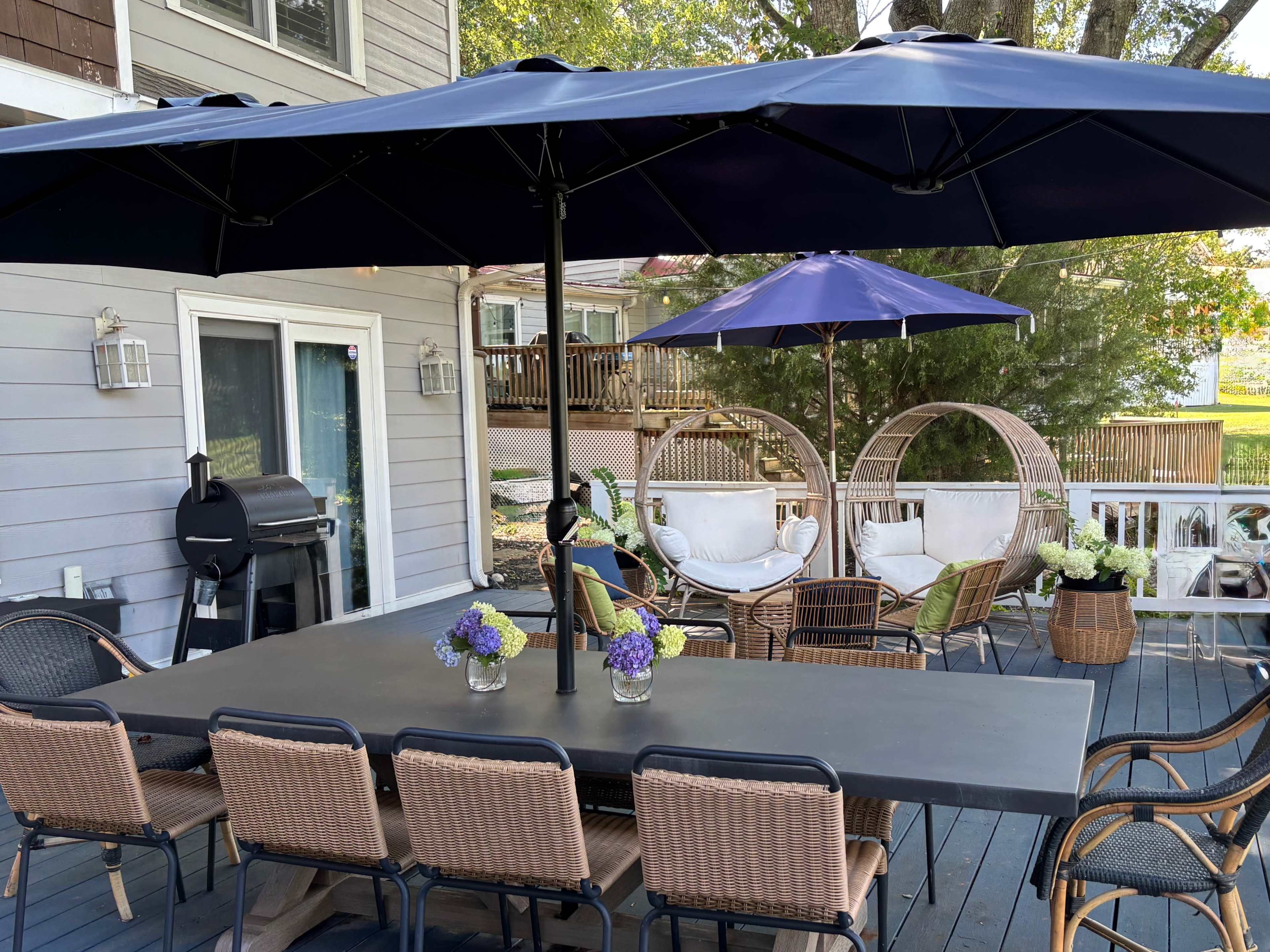 The image shows a patio area featuring a large dining table surrounded by chairs, two hanging chairs, and several umbrellas, with a barbecue grill positioned nearby.