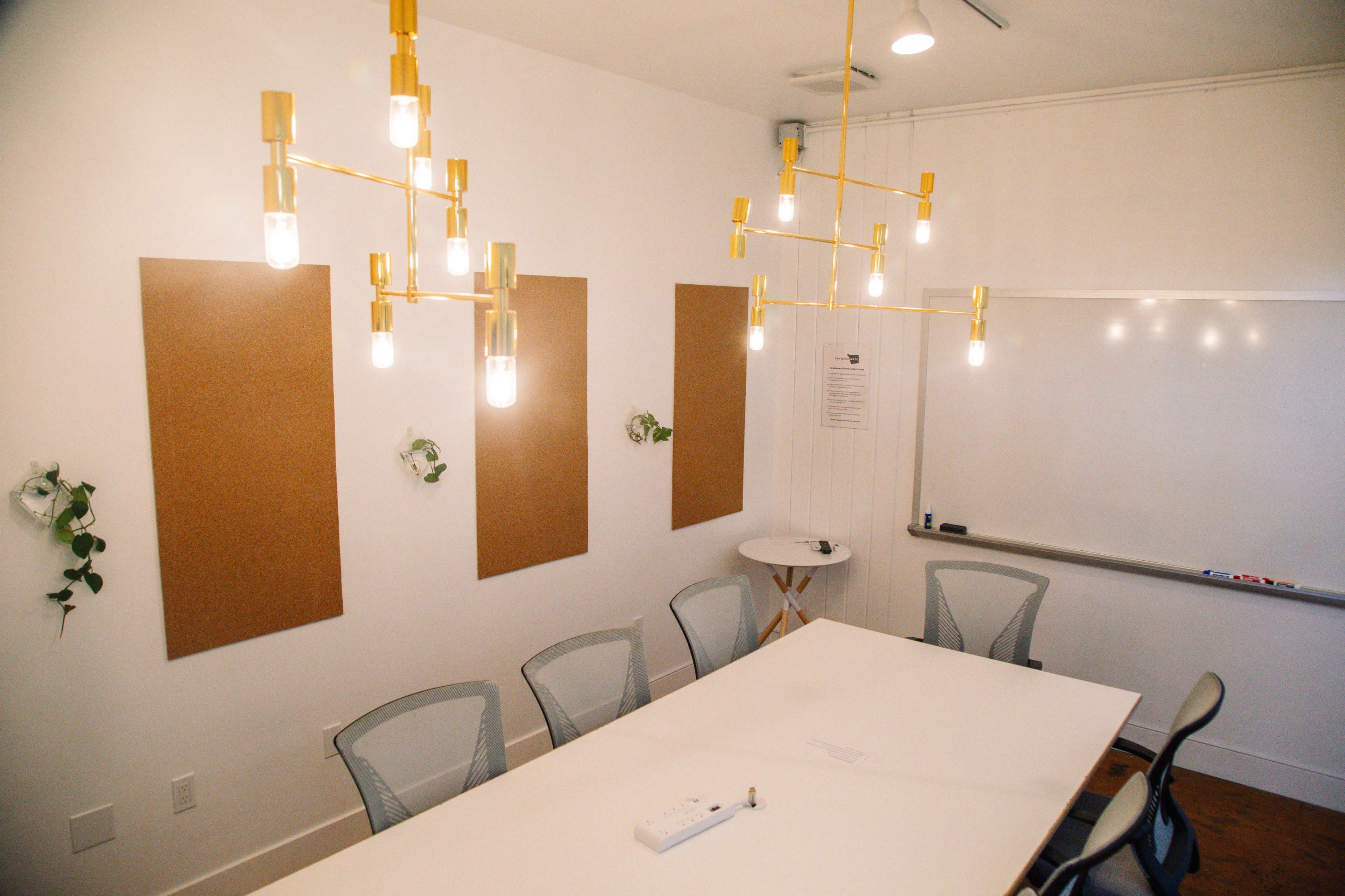 A modern conference room features a long white table, gold light fixtures, corkboard panels on the walls, and a whiteboard at one end.