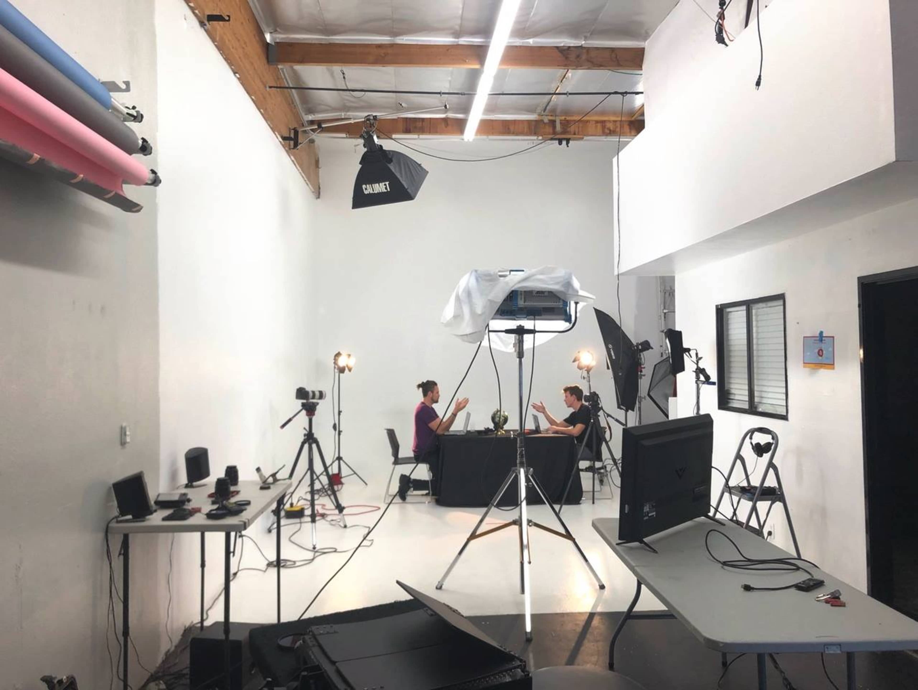 Two people sit at a table under lighting equipment in a minimalistic studio with various cameras and setup equipment around them.