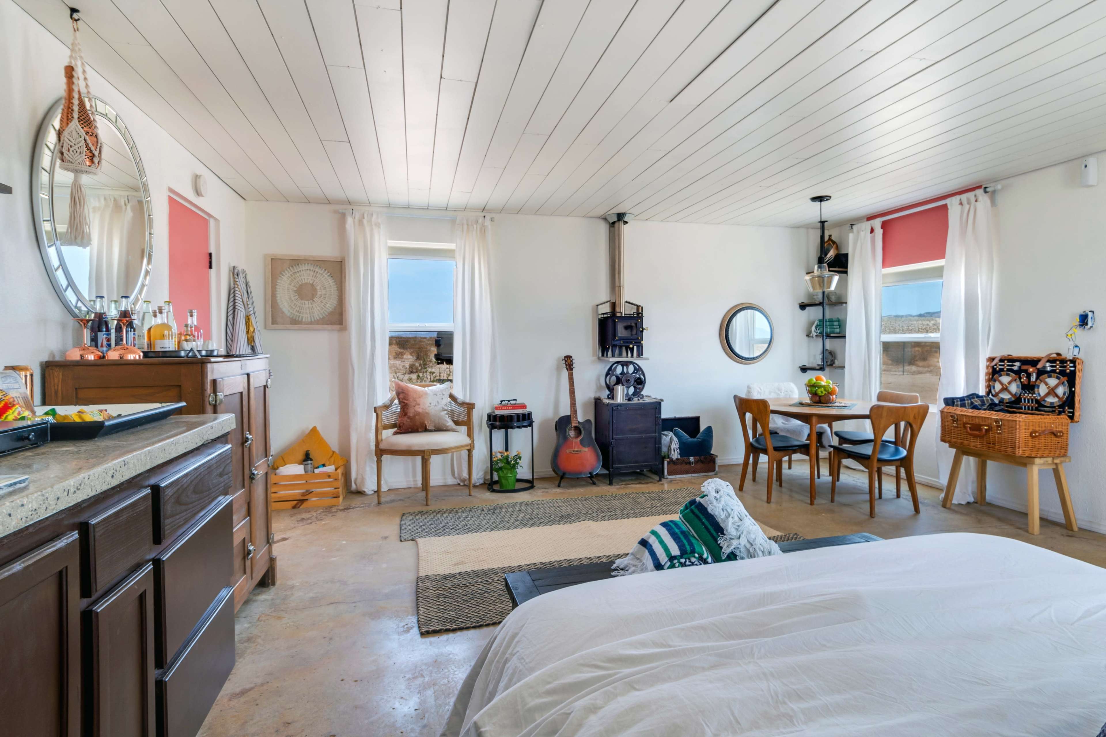 A compact room features a bed, sitting area, kitchen space, and dining table, with large windows overlooking a view.
