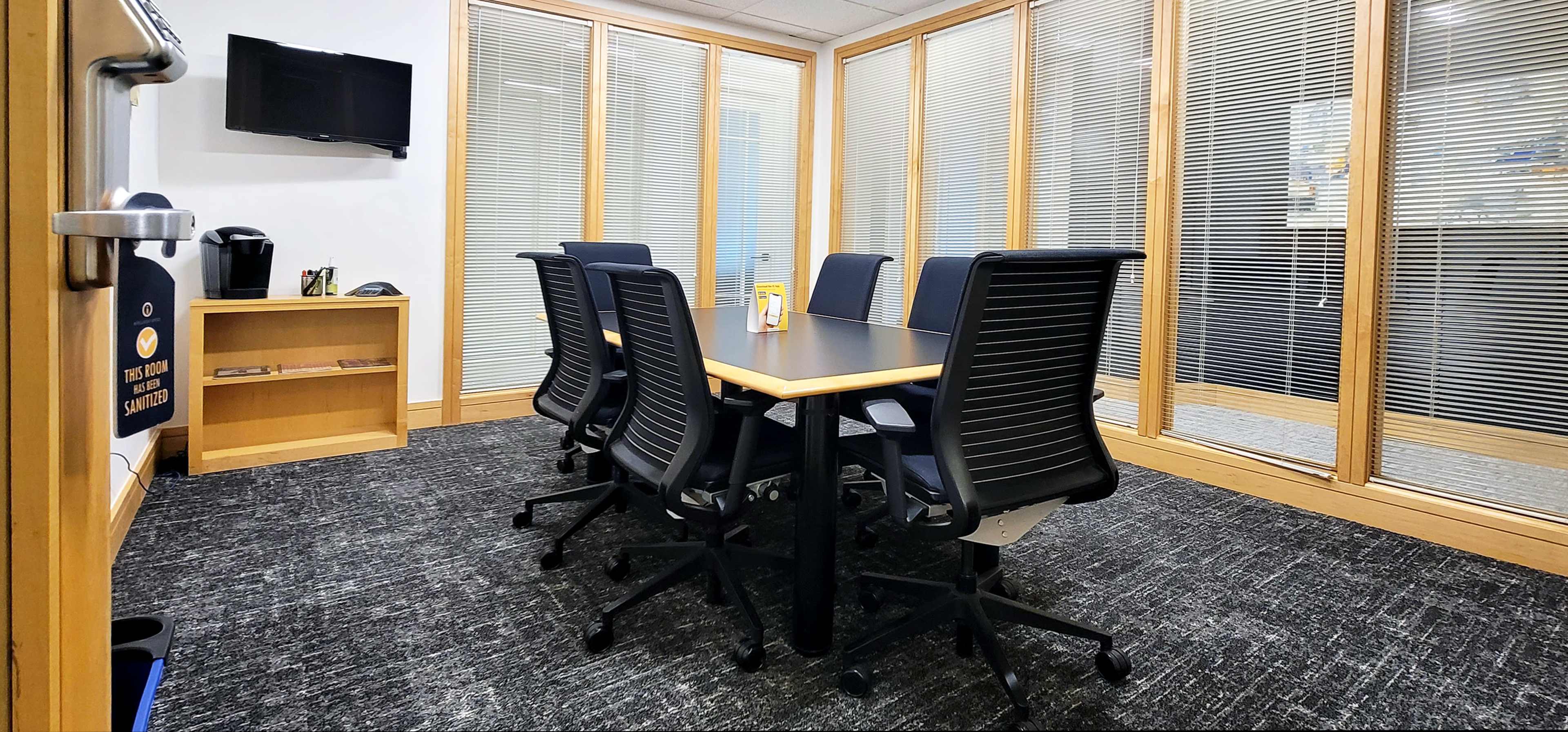 A conference room features a rectangular table surrounded by six black chairs, with glass-paneled walls and a television mounted on one side.