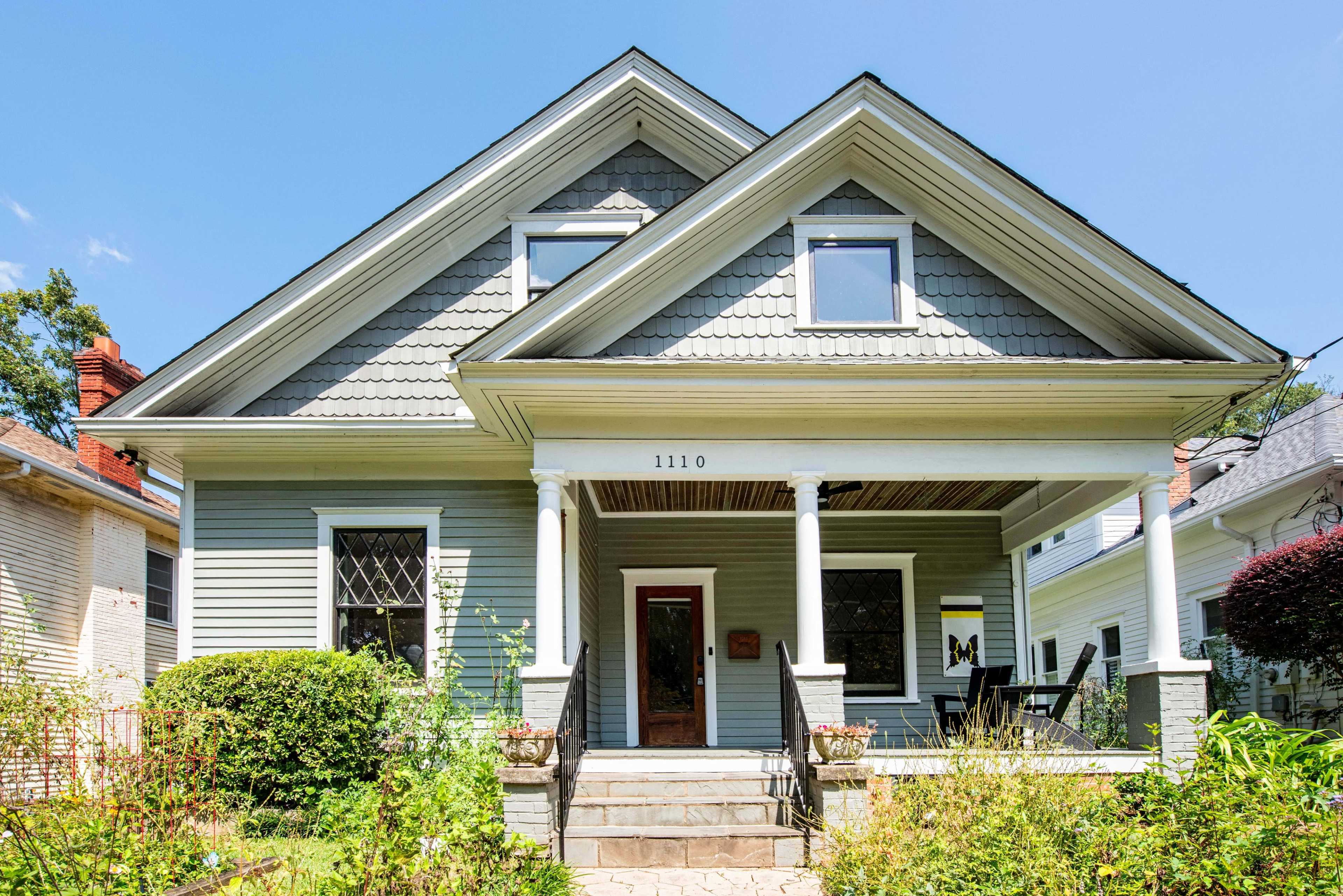 A light blue, two-story house features a prominent gable roof, a front porch with steps, and a well-maintained garden.