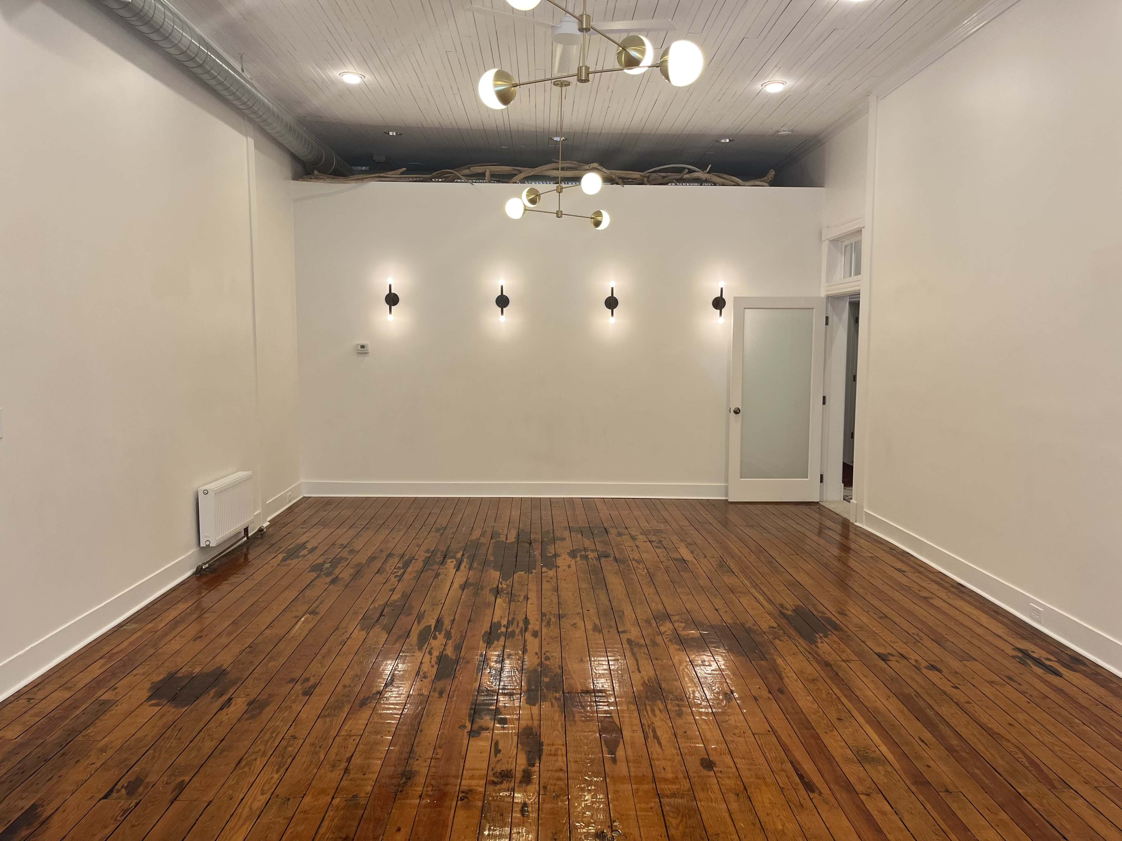 The image shows an empty room with wooden floors, white walls, and modern light fixtures hanging from the ceiling.