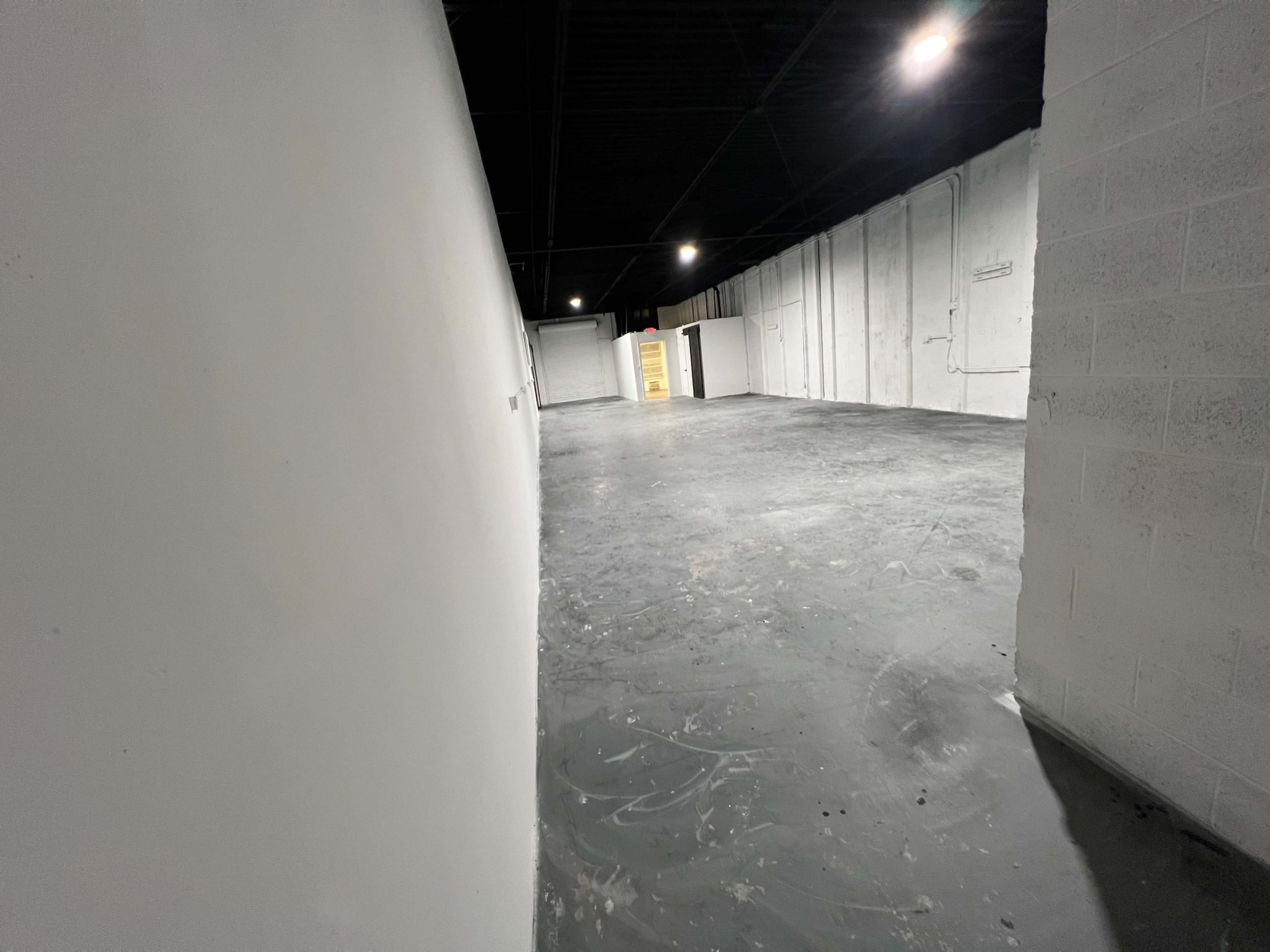 The image shows a large, empty room with a smooth, gray floor and white walls, illuminated by overhead lights.