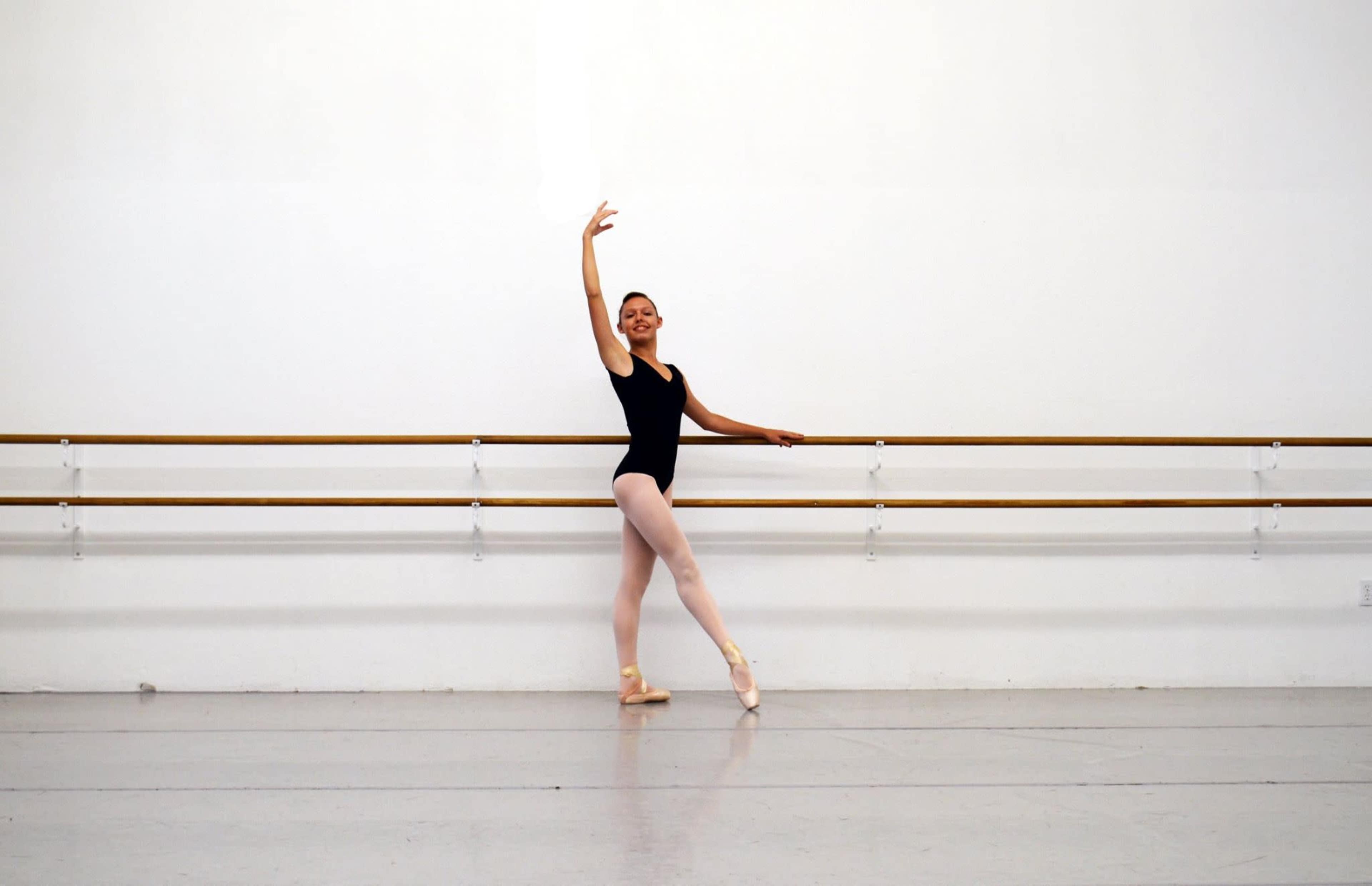 A ballet dancer poses gracefully in a studio with wooden ballet bars along the wall.