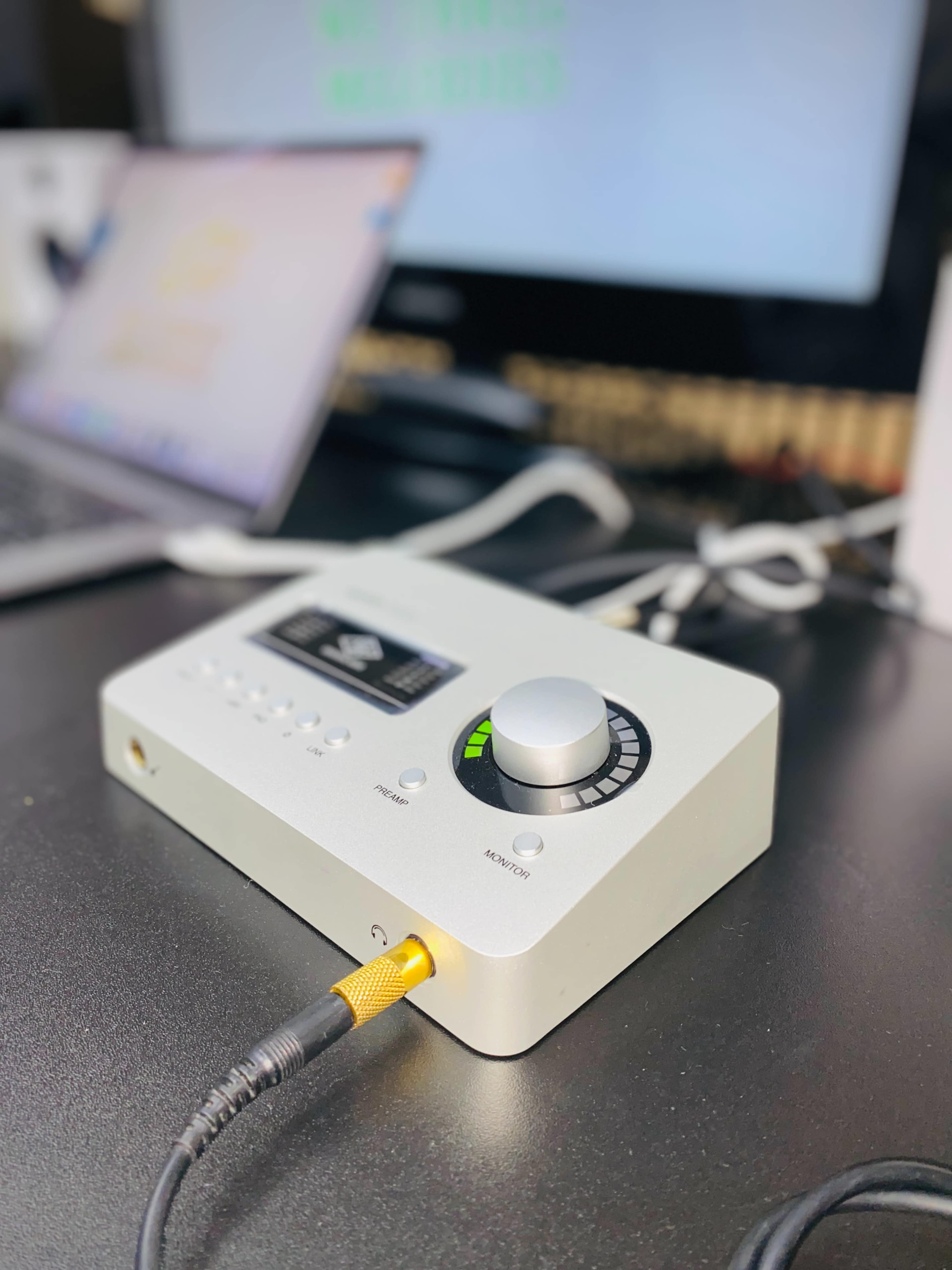 The image shows an audio interface with a large volume knob, connected to a computer and other cables on a desk.