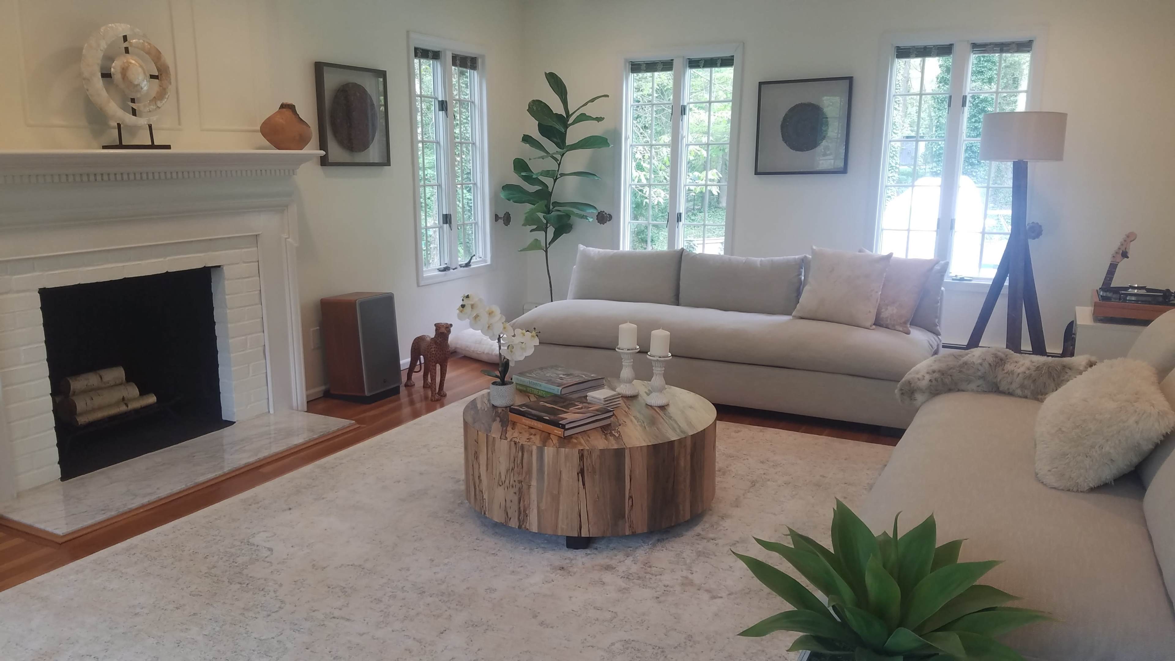 A bright living room features a light gray sectional sofa, a round wooden coffee table, a stone fireplace, and large windows allowing natural light.