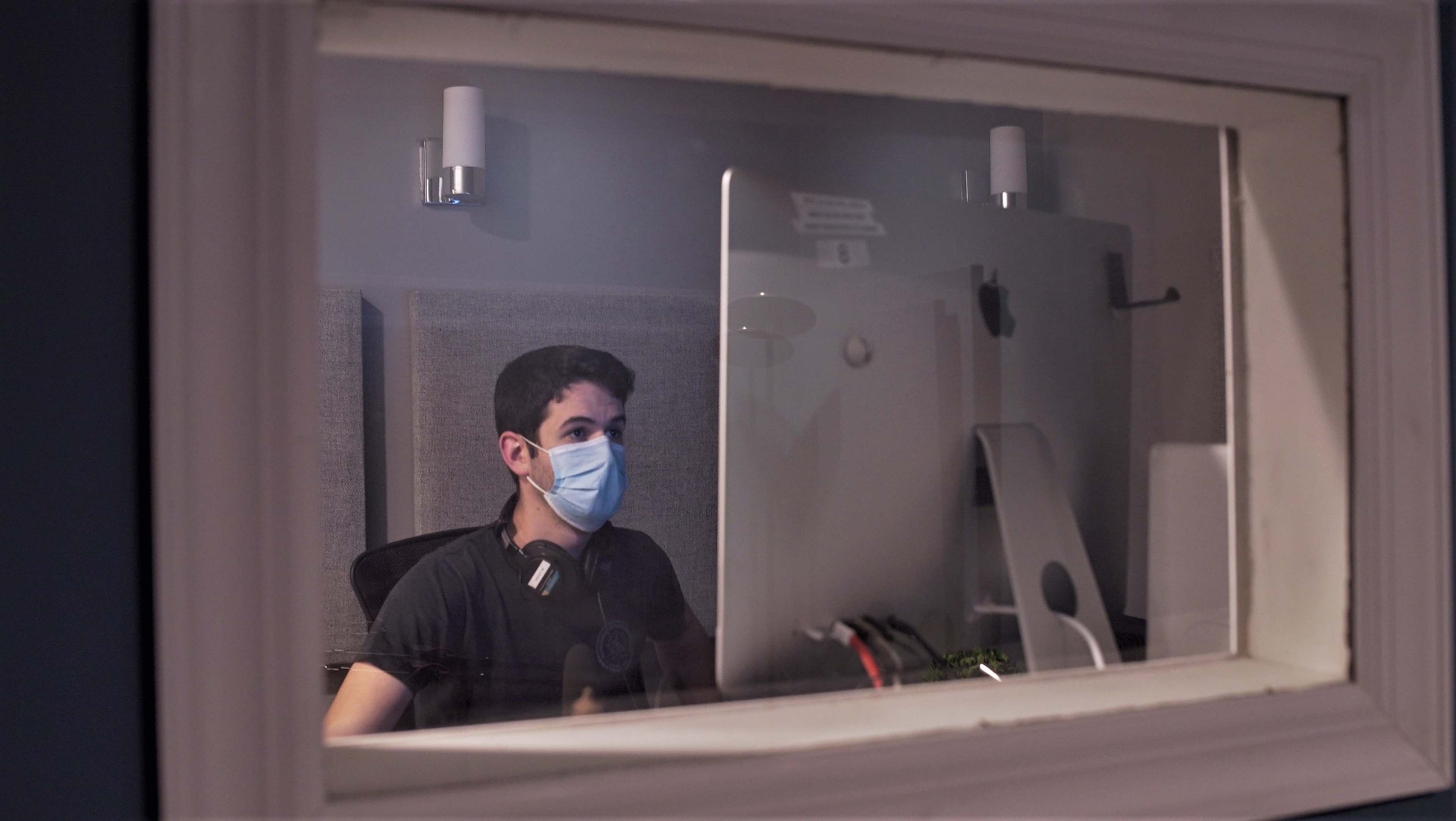A young man wearing a face mask sits at a desk in front of a computer, with headphones around his neck, in a room visible through a window.