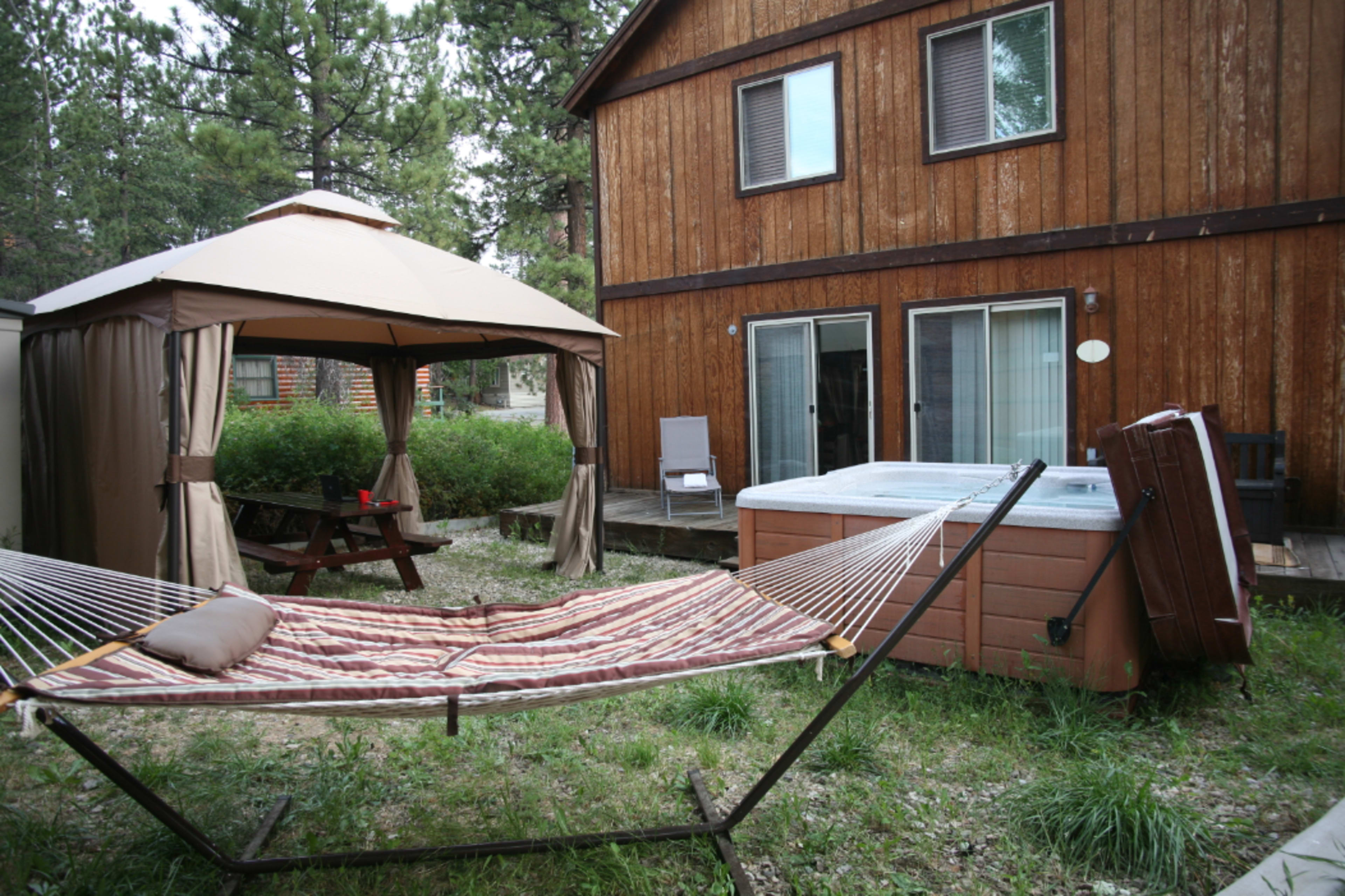 A backyard area features a hammock, a gazebo, a picnic table, and a hot tub next to a wooden house.