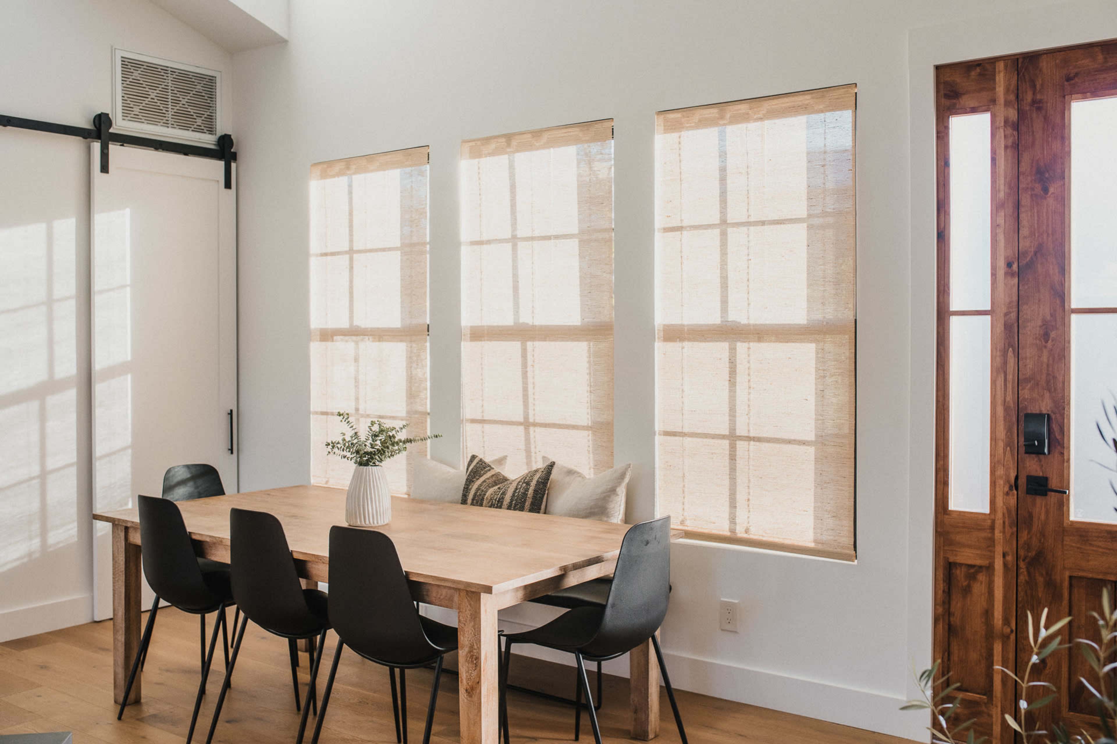 A wooden dining table is surrounded by black chairs in a bright room with large windows covered by bamboo shades.