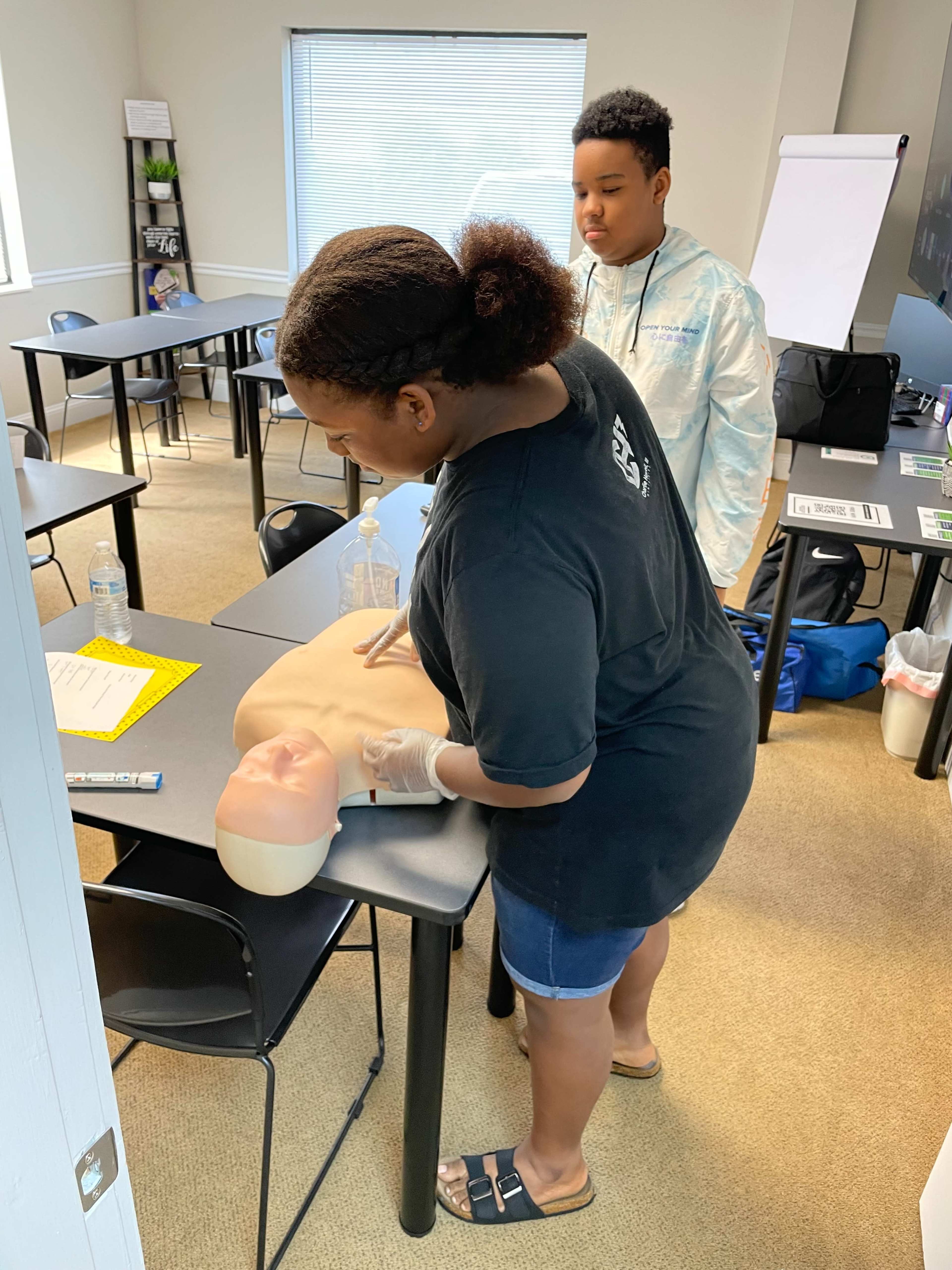 A woman practices CPR on a manikin while a young boy observes in a classroom setting.