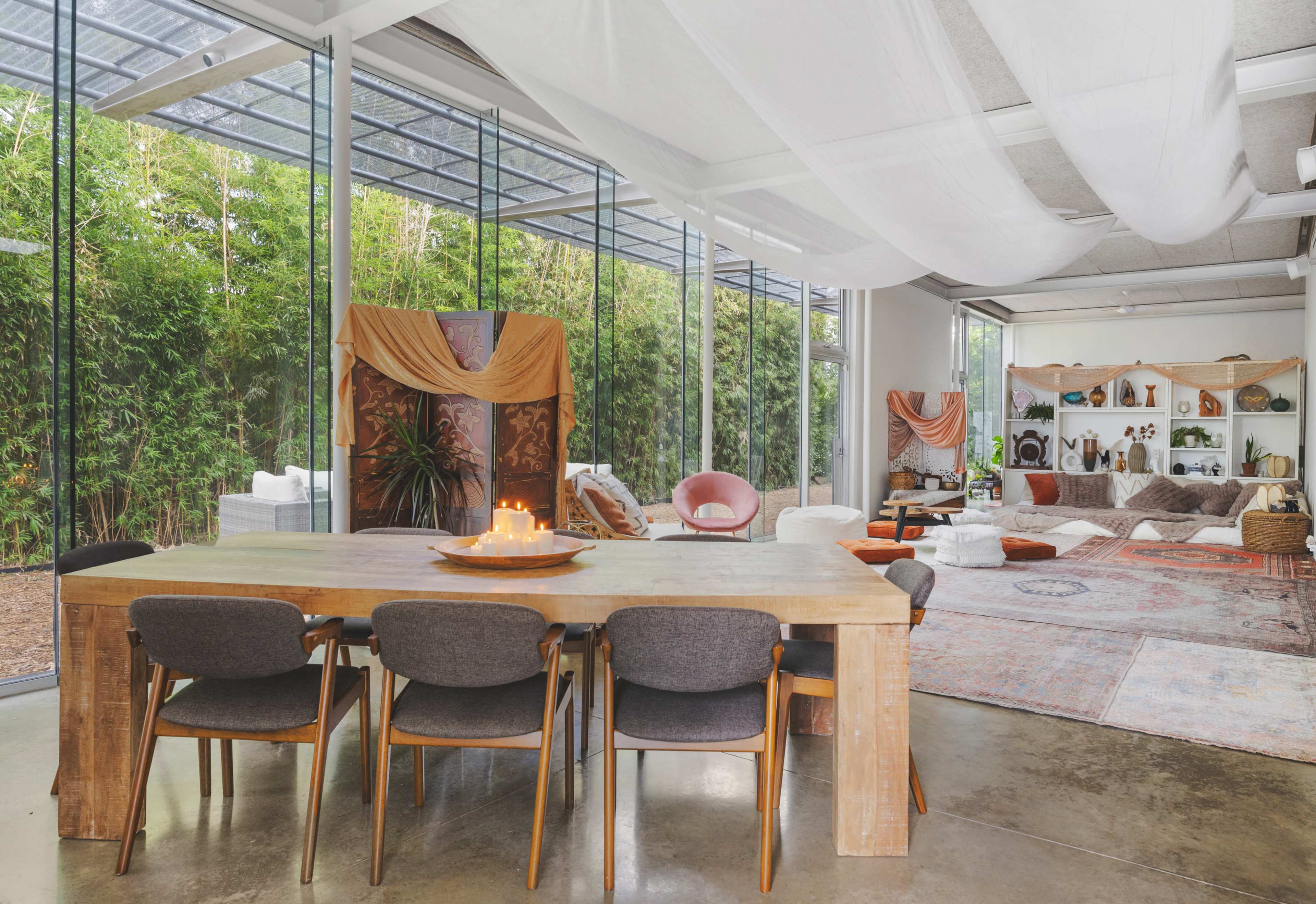 A spacious, modern living area features a large wooden dining table surrounded by gray chairs, with greenery visible through glass walls and various seating arrangements in a cozy, decorated corner.