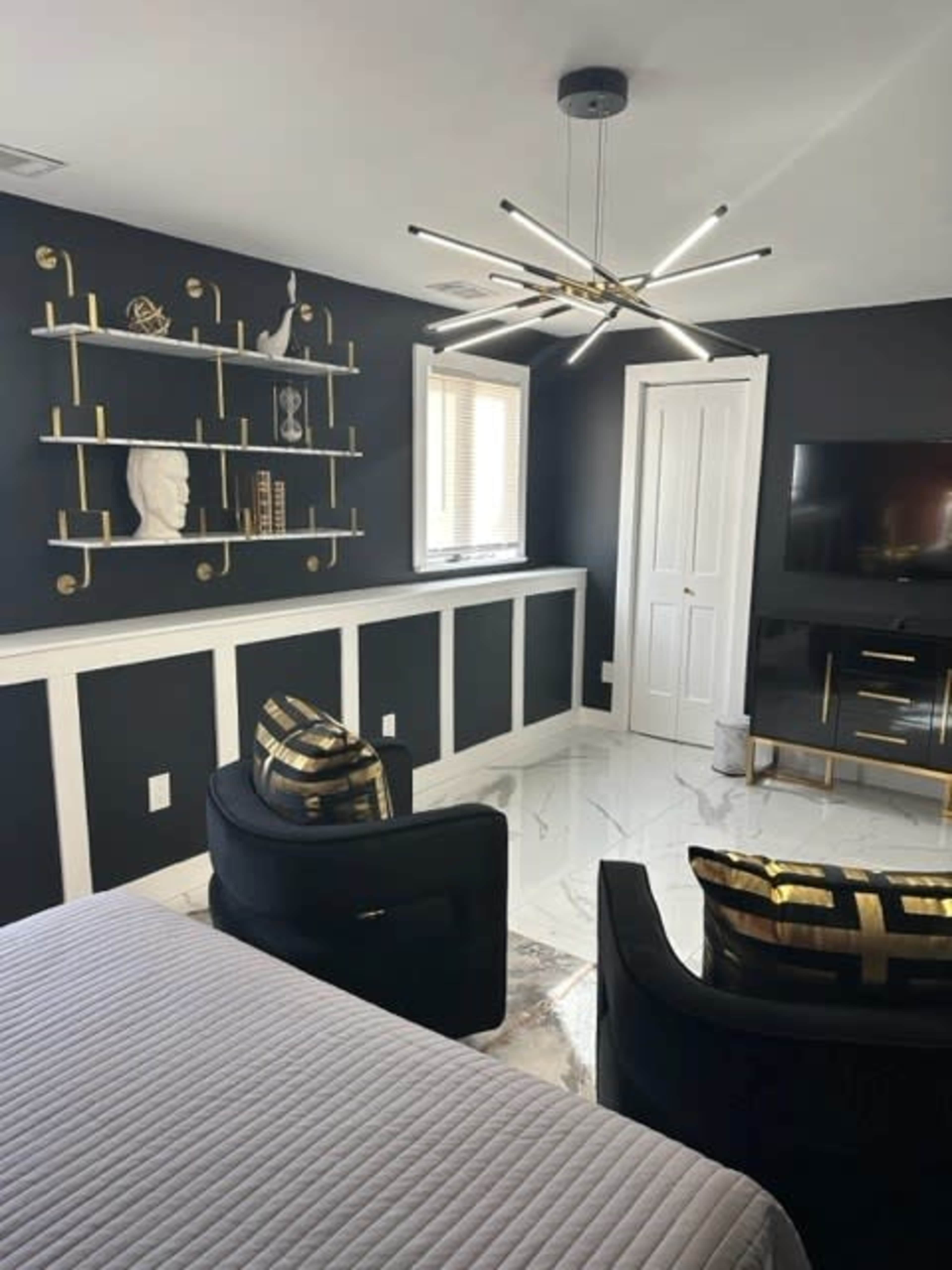 A modern bedroom featuring black walls, white trim, a bed with a gray quilt, two black chairs, and a mounted television.