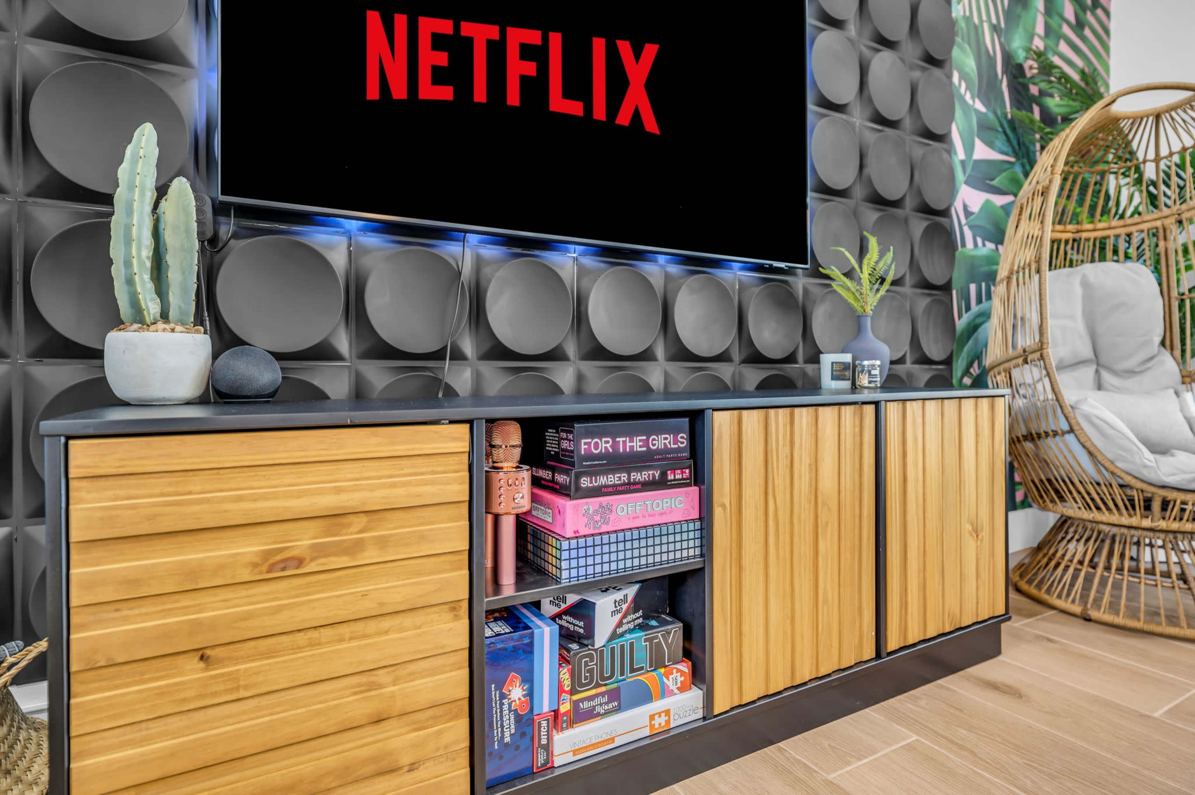 A wooden entertainment unit with games and decorative items sits below a large TV displaying the Netflix logo, against a backdrop of textured wall panels and plants.