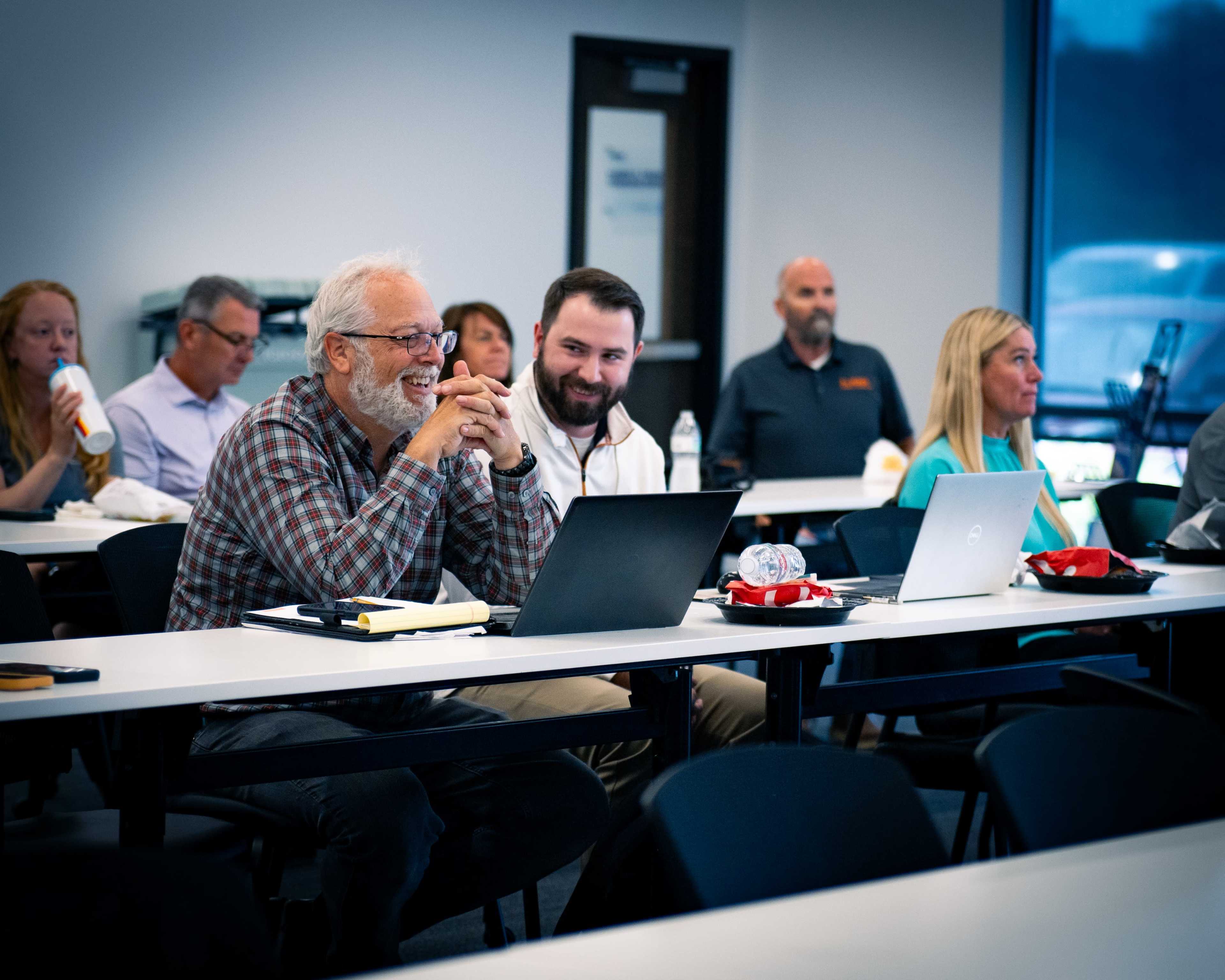 A group of people sits in a classroom setting, engaged with laptops open on desks.