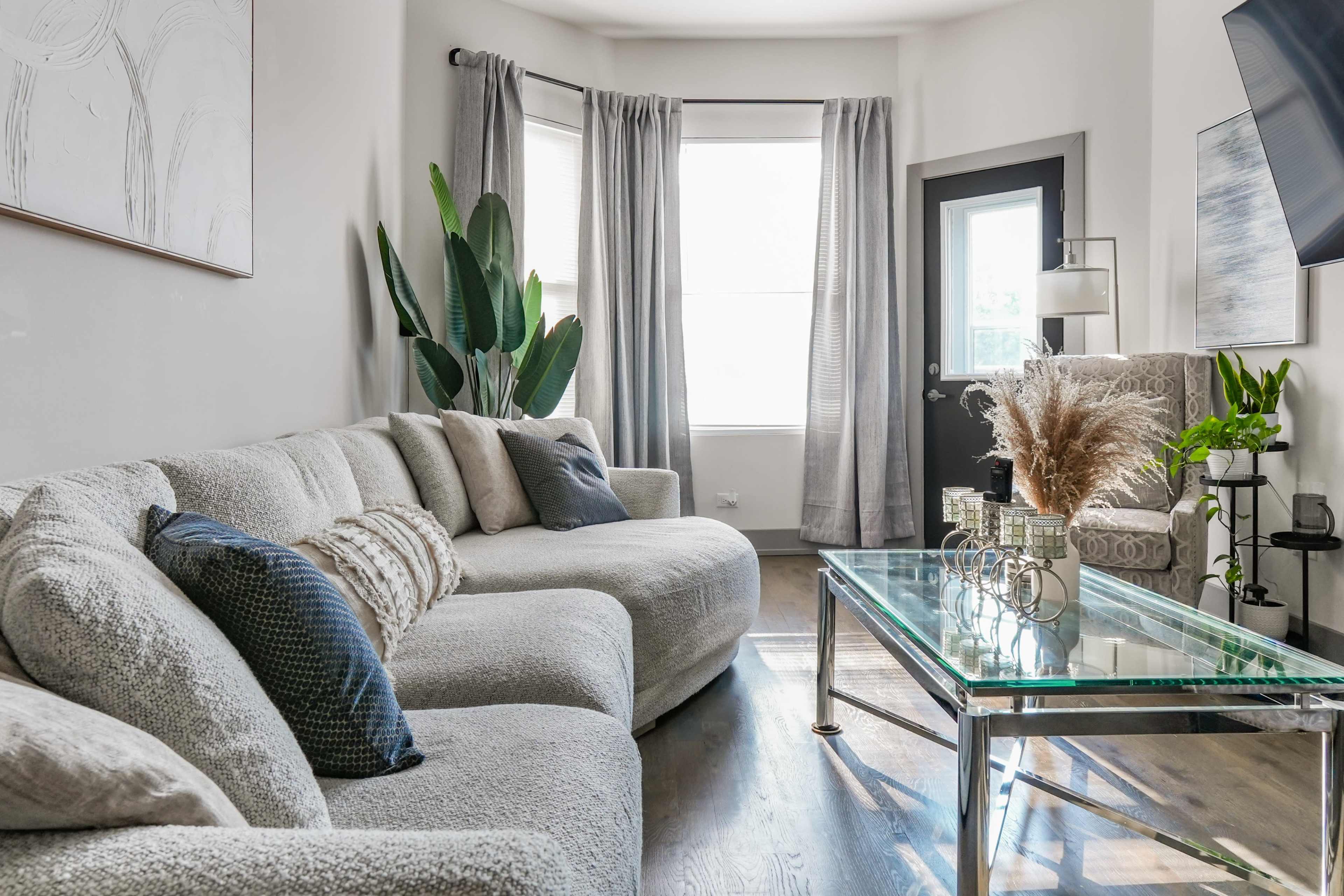 A cozy living room features a large, light-colored sectional sofa, a glass coffee table, and potted plants, with natural light streaming in through a window.