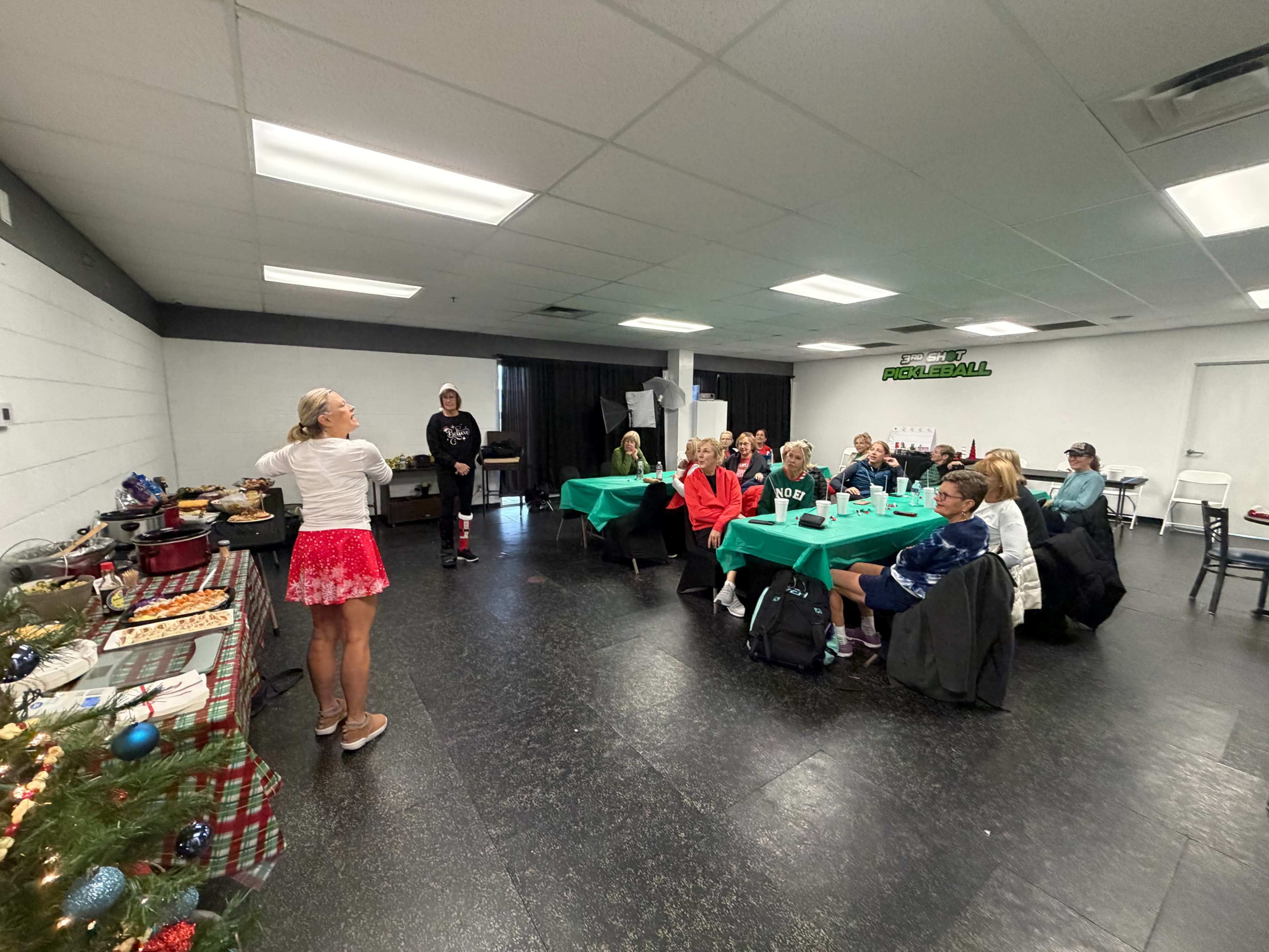 A group of people seated at tables in a room decorated for a gathering watch a woman speaking in front of a table filled with food.