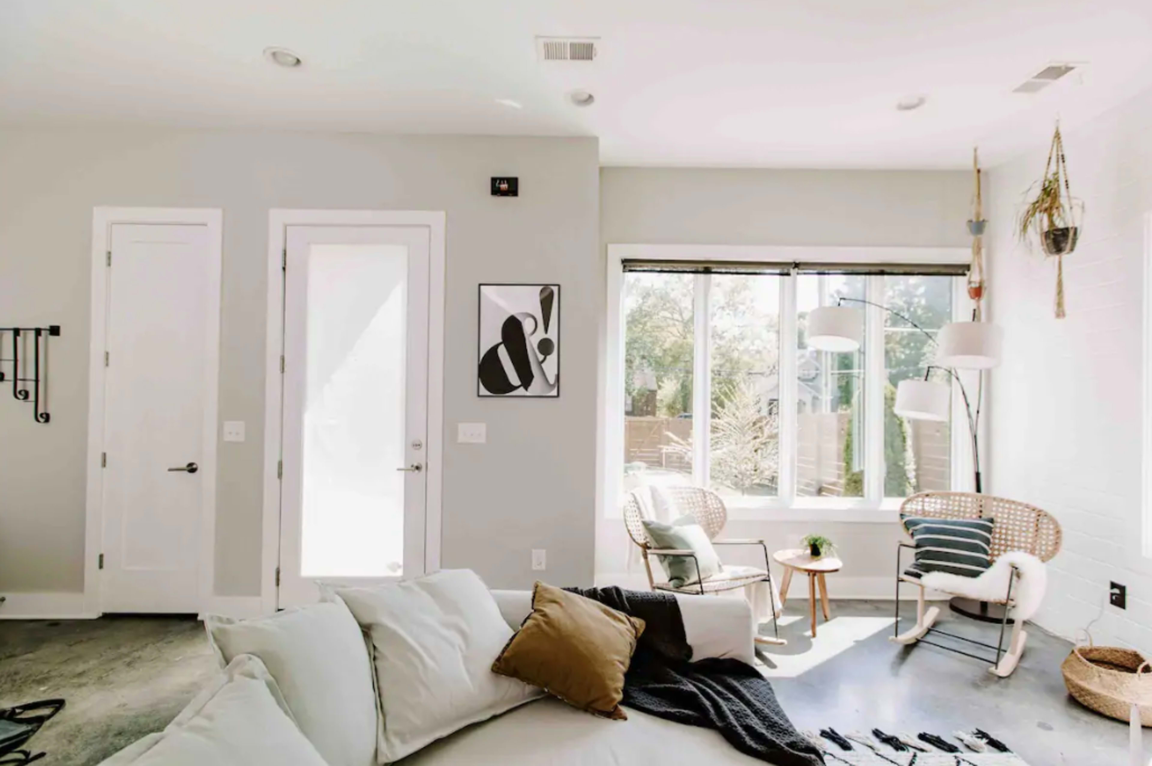The image shows a bright, modern living room with a white sofa, a framed artwork on the wall, and a sunlit corner featuring two chairs and a large window.