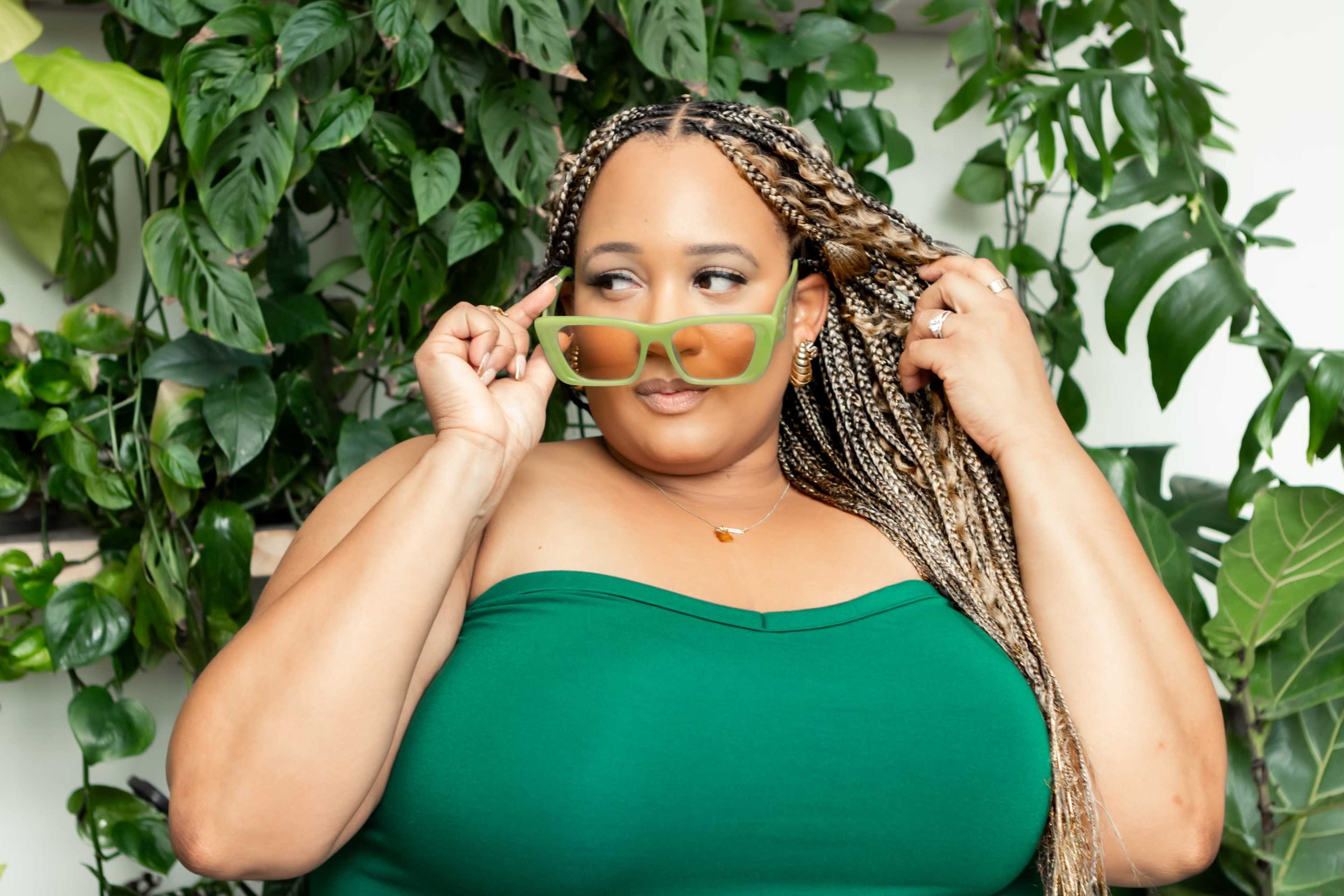 A woman with long braided hair wearing green sunglasses poses in front of a backdrop of lush green plants.