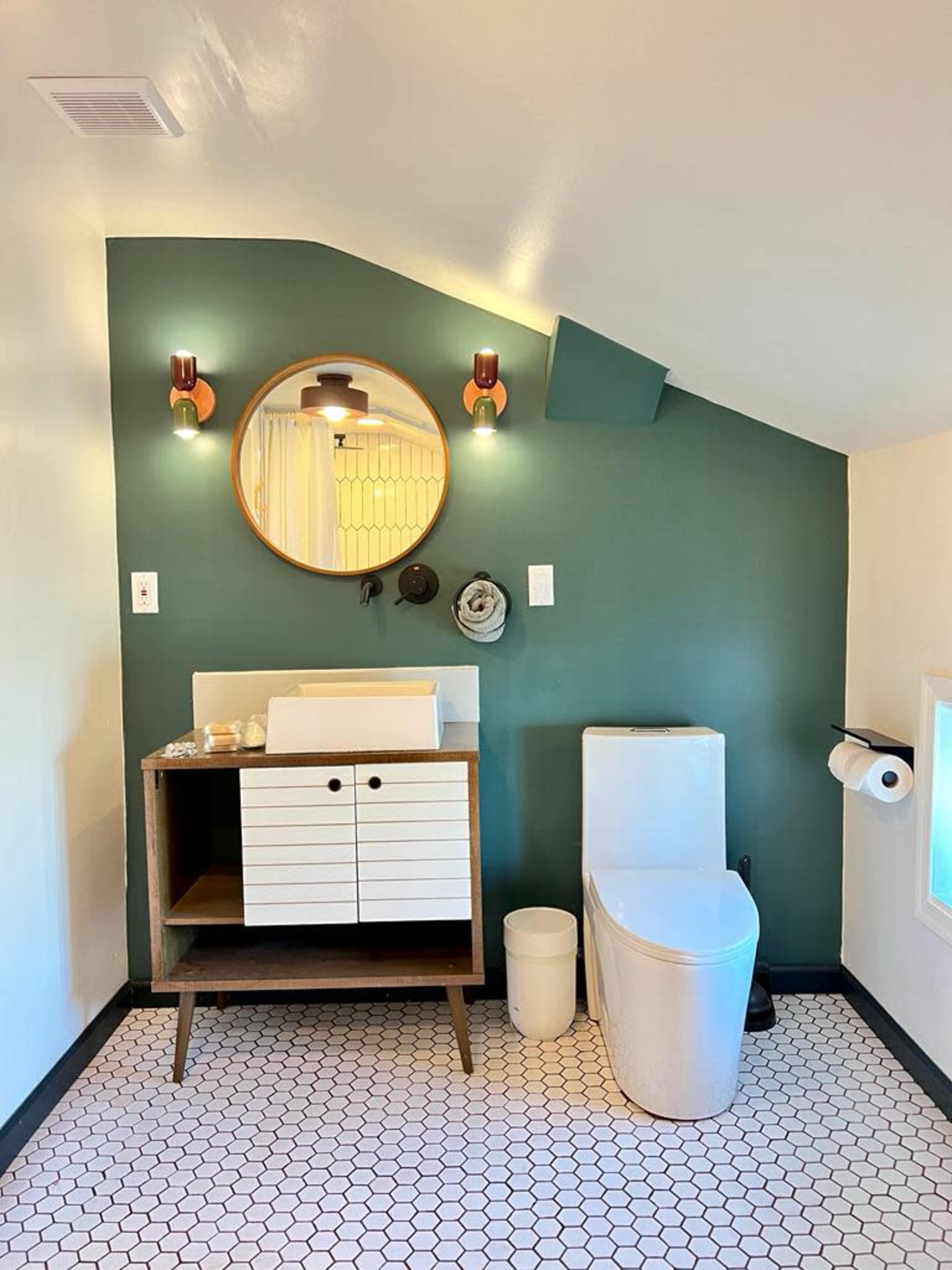 A bathroom featuring a green wall, a circular mirror, a white toilet, and a wooden vanity with white sliding drawers, all set against a hexagonal tiled floor.