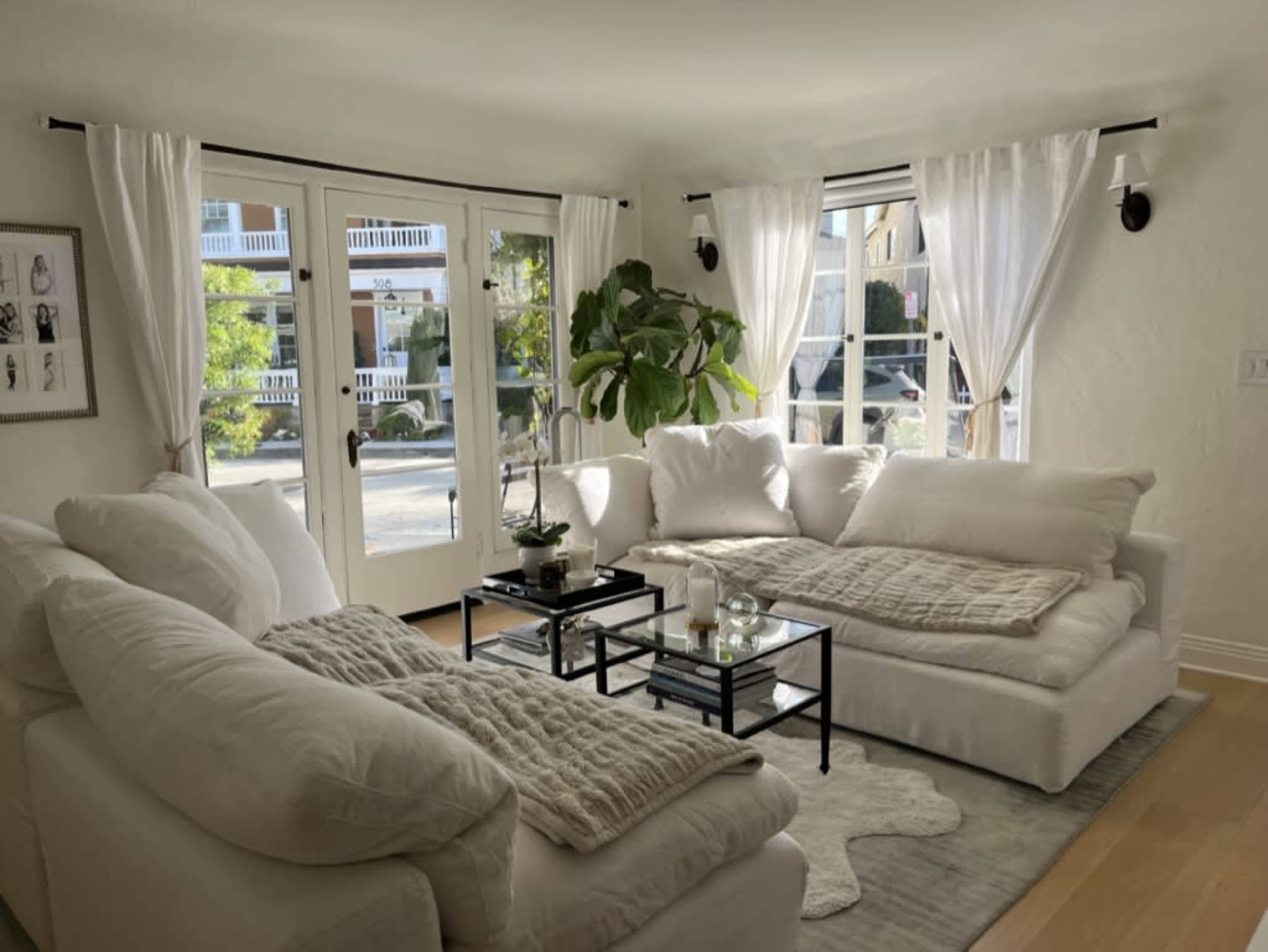 A bright living room with large windows, white furniture, and a small coffee table adorned with candles and a potted plant.