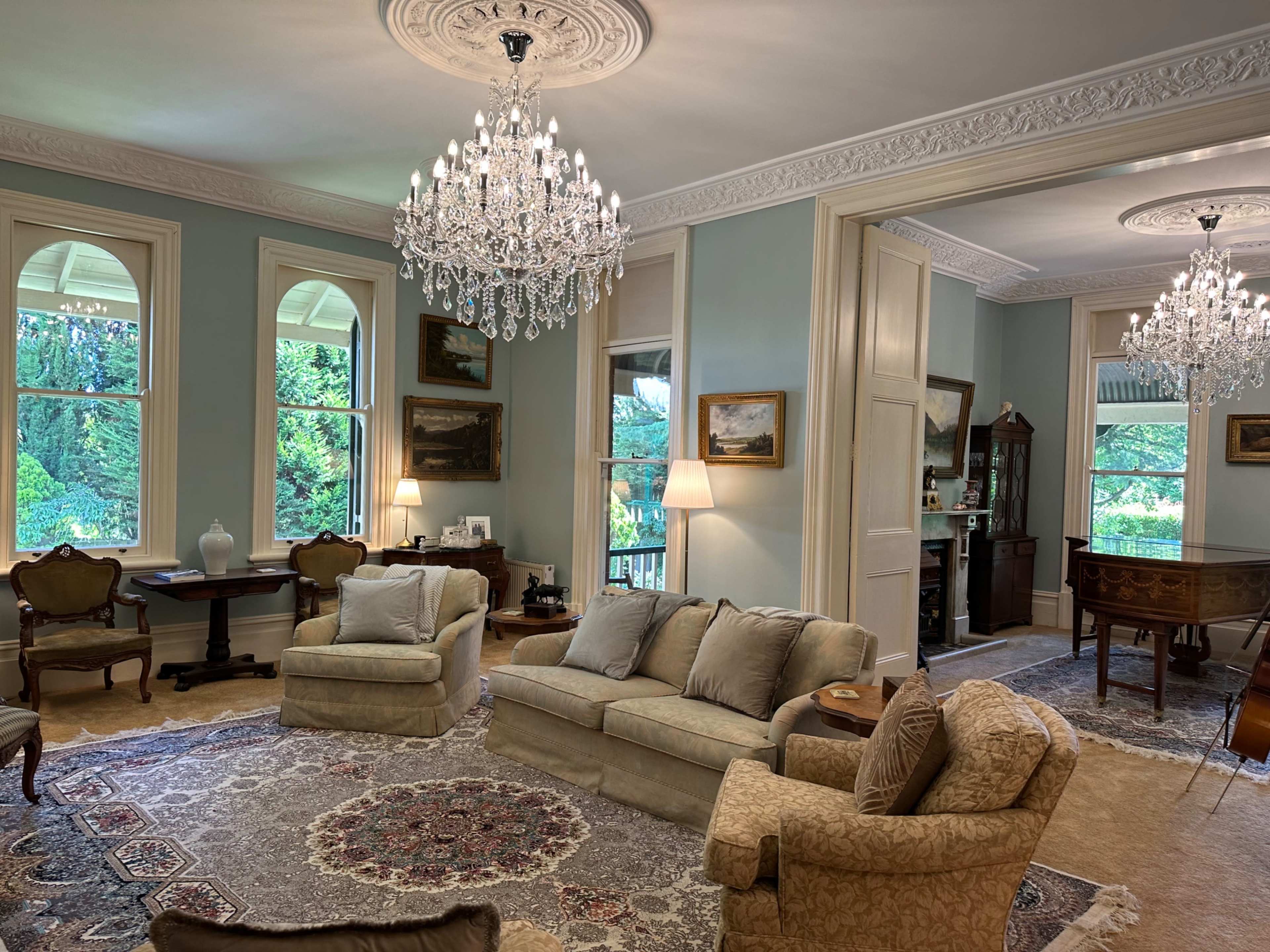 The image shows a spacious living room with two chandeliers, elegant furniture, and large windows overlooking greenery.