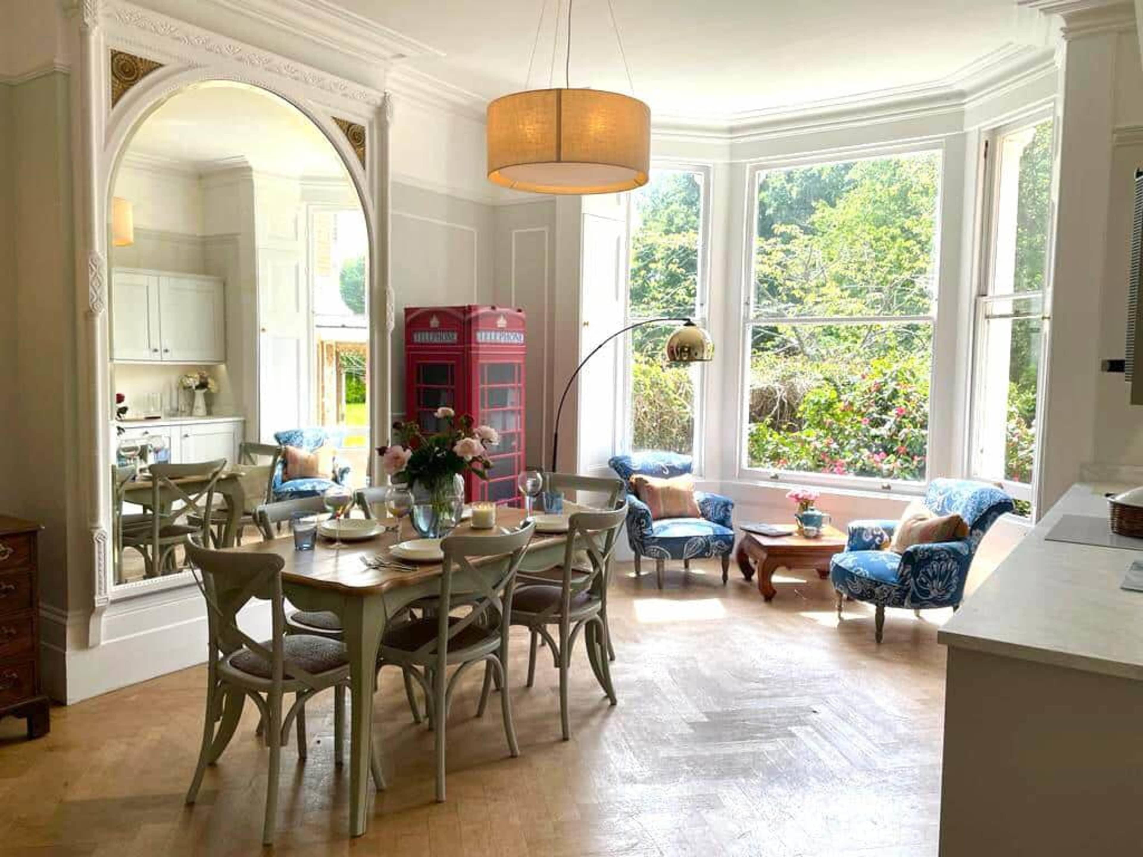 A dining area features a circular table surrounded by chairs, with a large window overlooking a garden and a red phone booth in the corner.