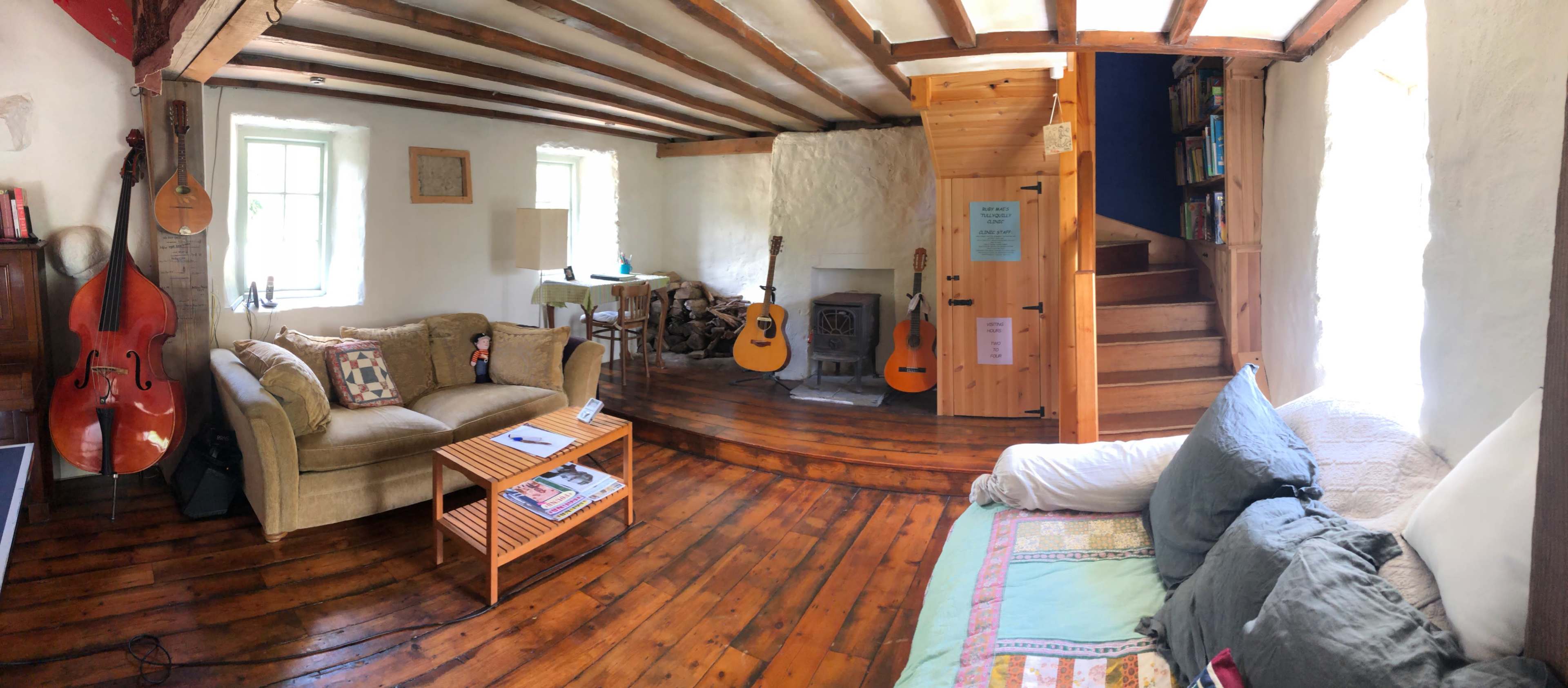 The image shows a cozy living room with wooden floors, a wood-burning stove, and various musical instruments, including a cello and guitars, arranged throughout the space.