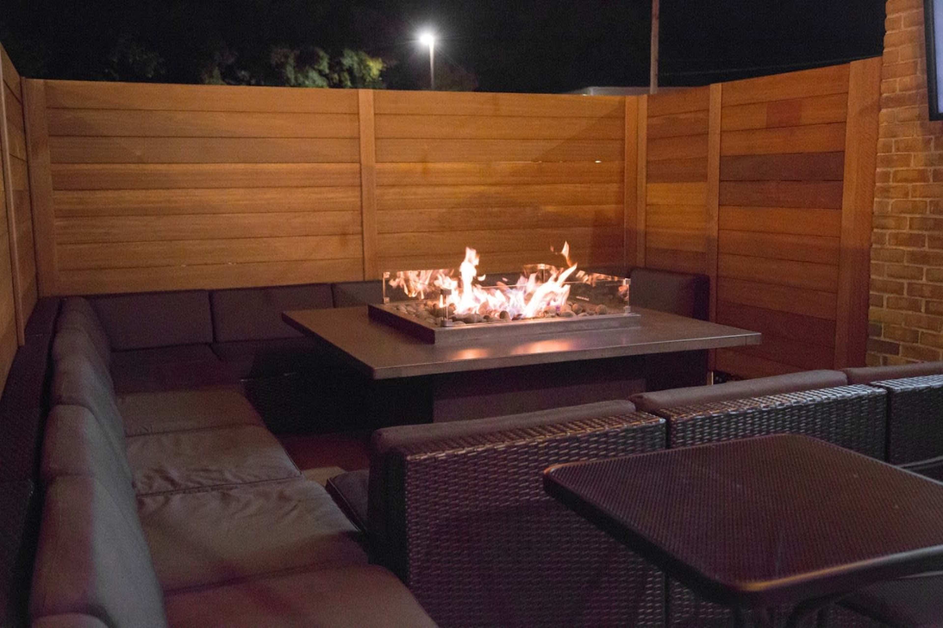 A fire pit surrounded by cushioned seating is situated on a patio enclosed by wooden panels.