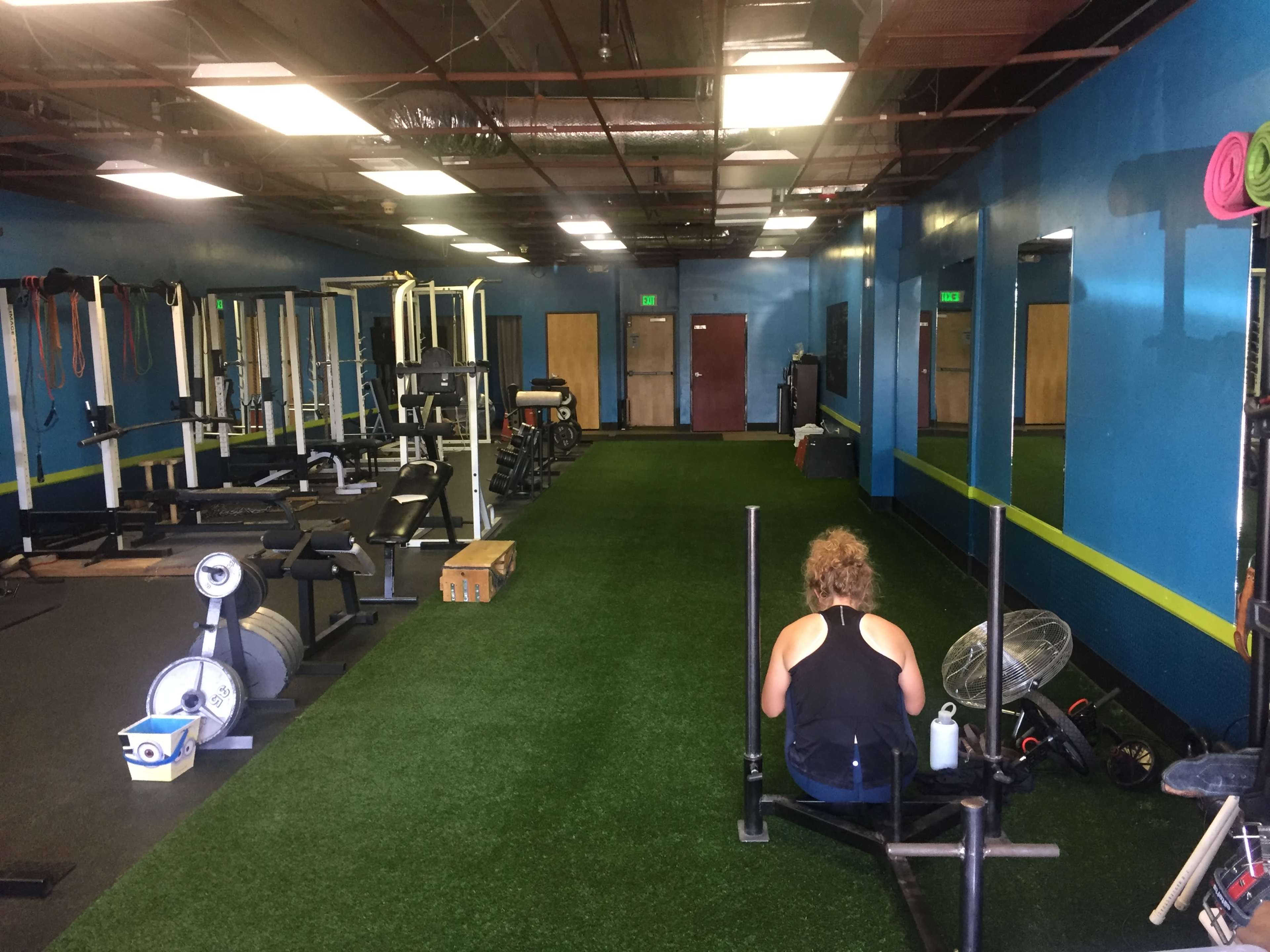 A person sits on a rowing machine in a gym with various exercise equipment and green turf flooring.