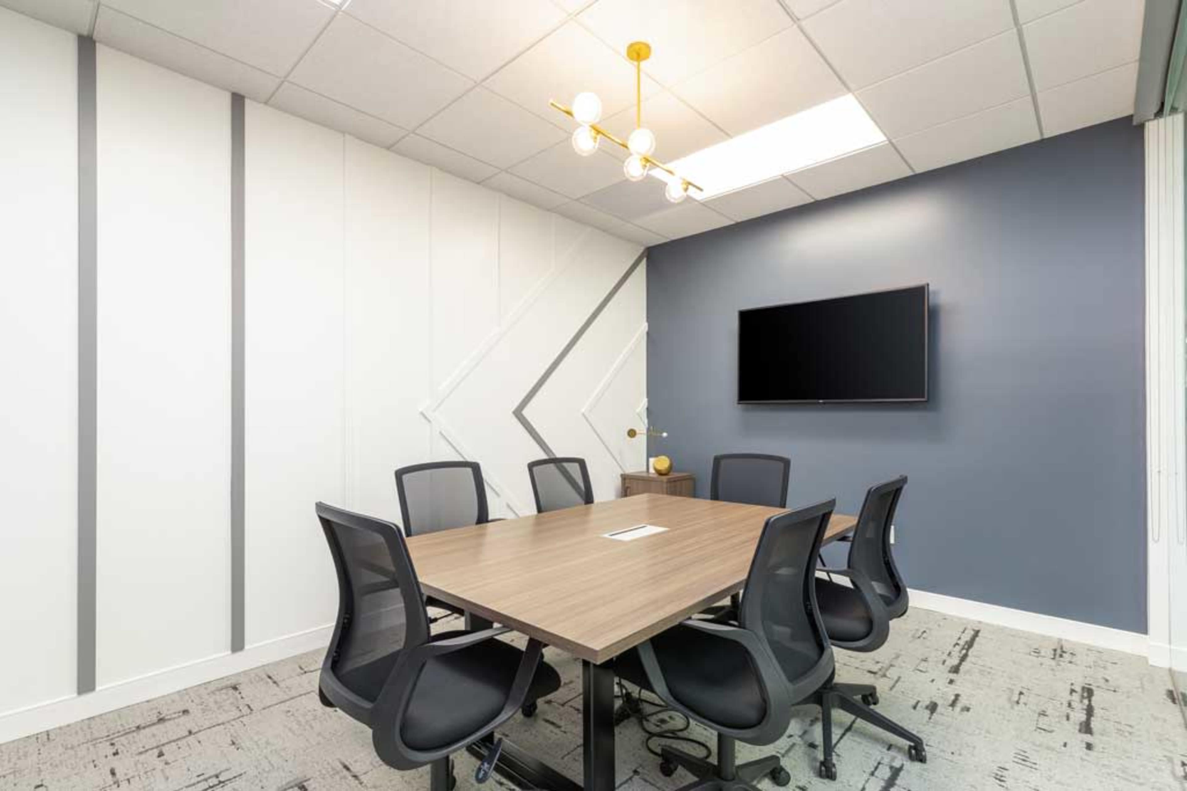 A modern conference room features a rectangular table surrounded by black office chairs, with a wall-mounted TV and decorative lighting.