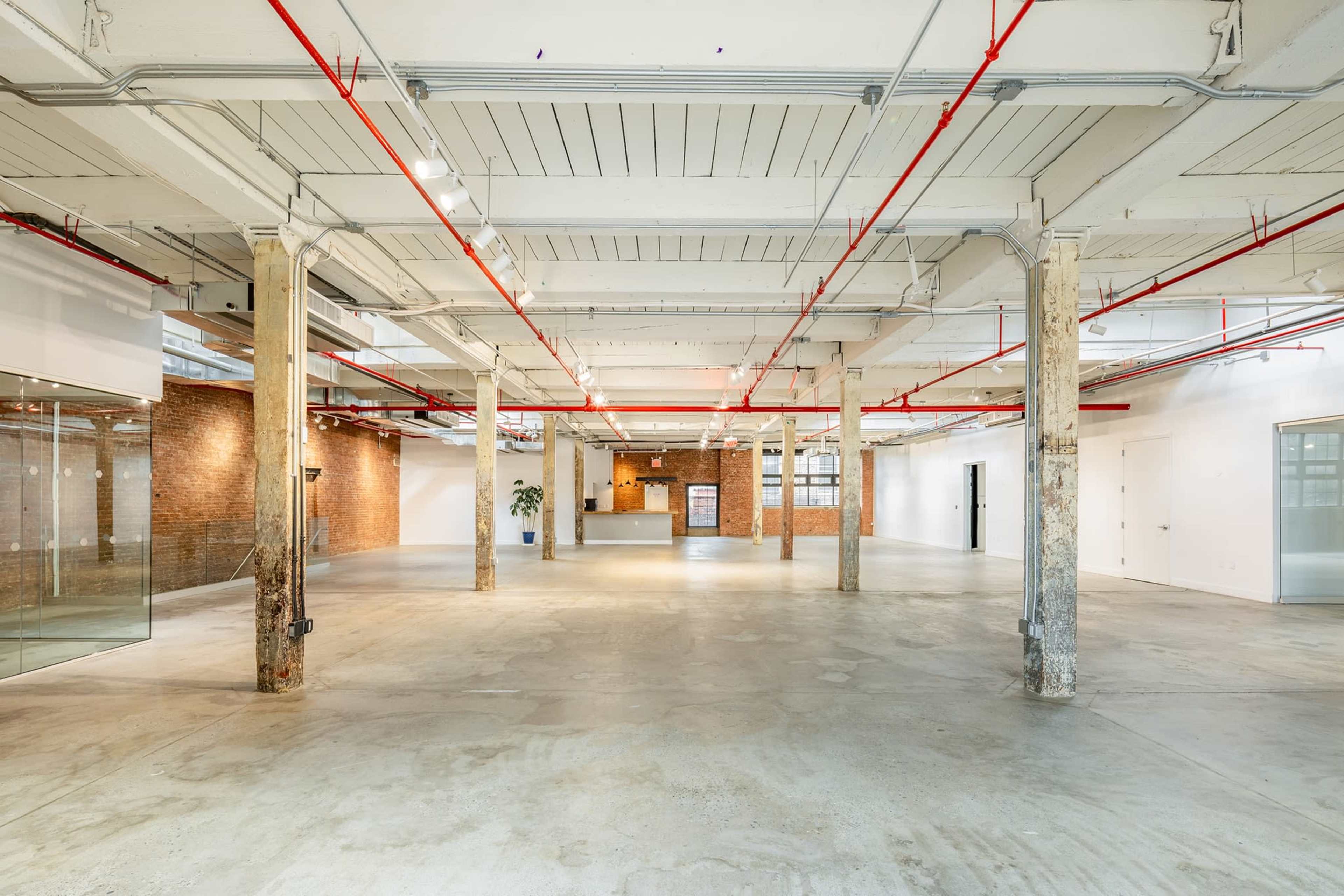 A spacious, unfinished industrial-style interior features exposed brick walls, concrete floors, and red ceiling pipes.