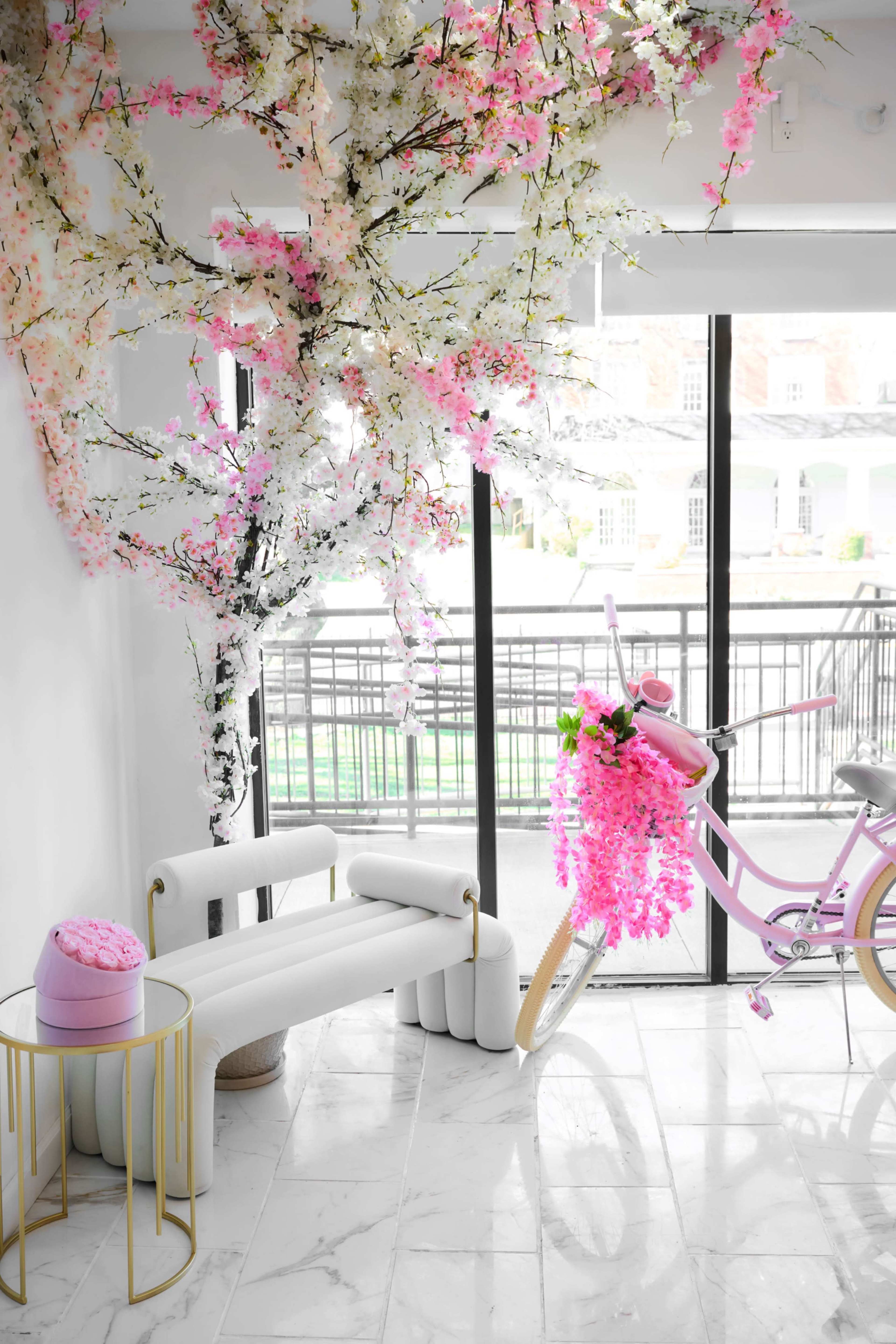 The scene features a well-lit interior with a white chaise lounge, a pink bicycle adorned with flowers, and artificial cherry blossoms hanging from the ceiling.