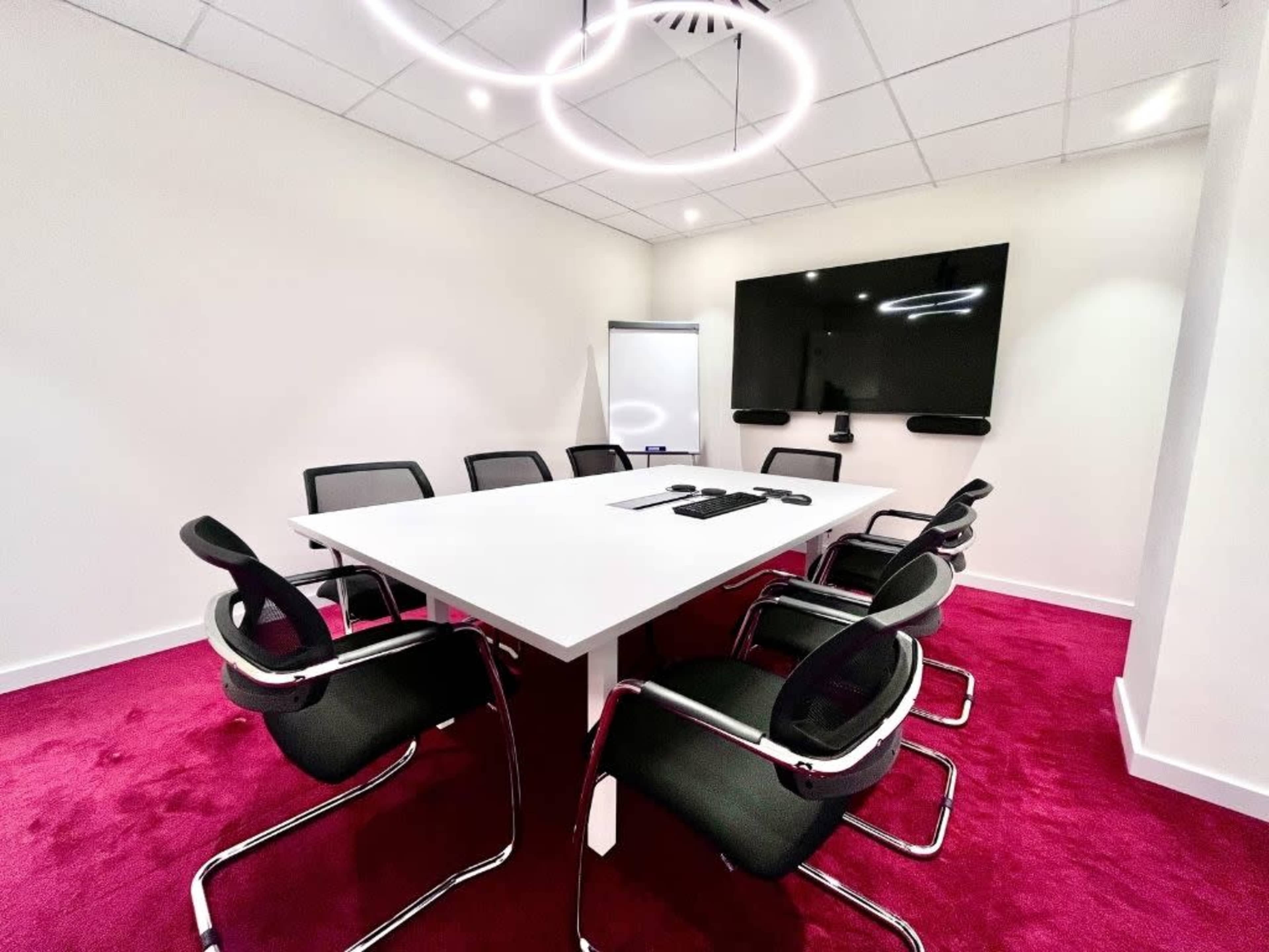 A modern conference room features a large white table surrounded by black chairs, with a wall-mounted screen and a flip chart in the background.