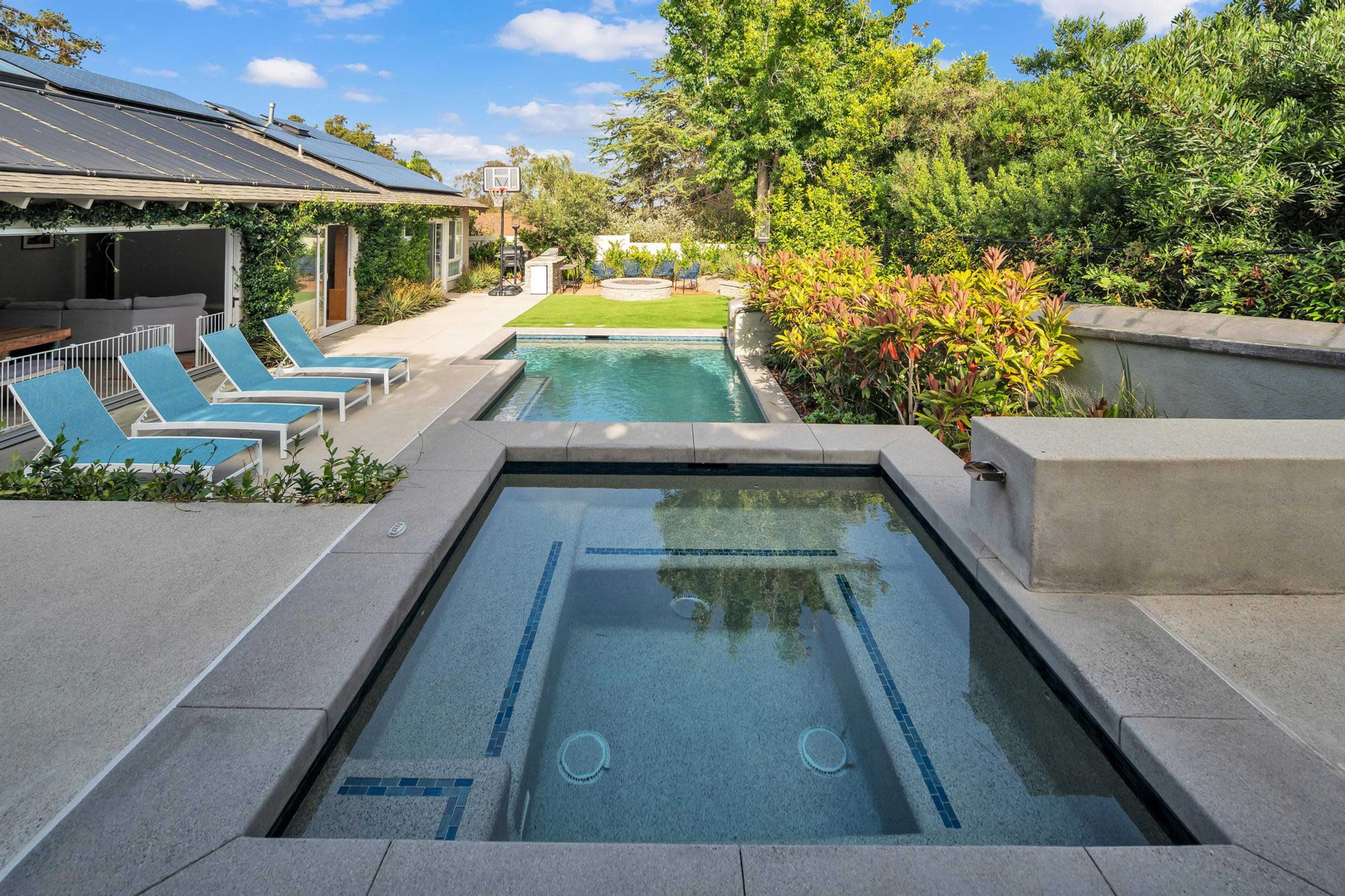 The image shows a landscaped backyard featuring a swimming pool, a hot tub, and lounge chairs under blue skies.